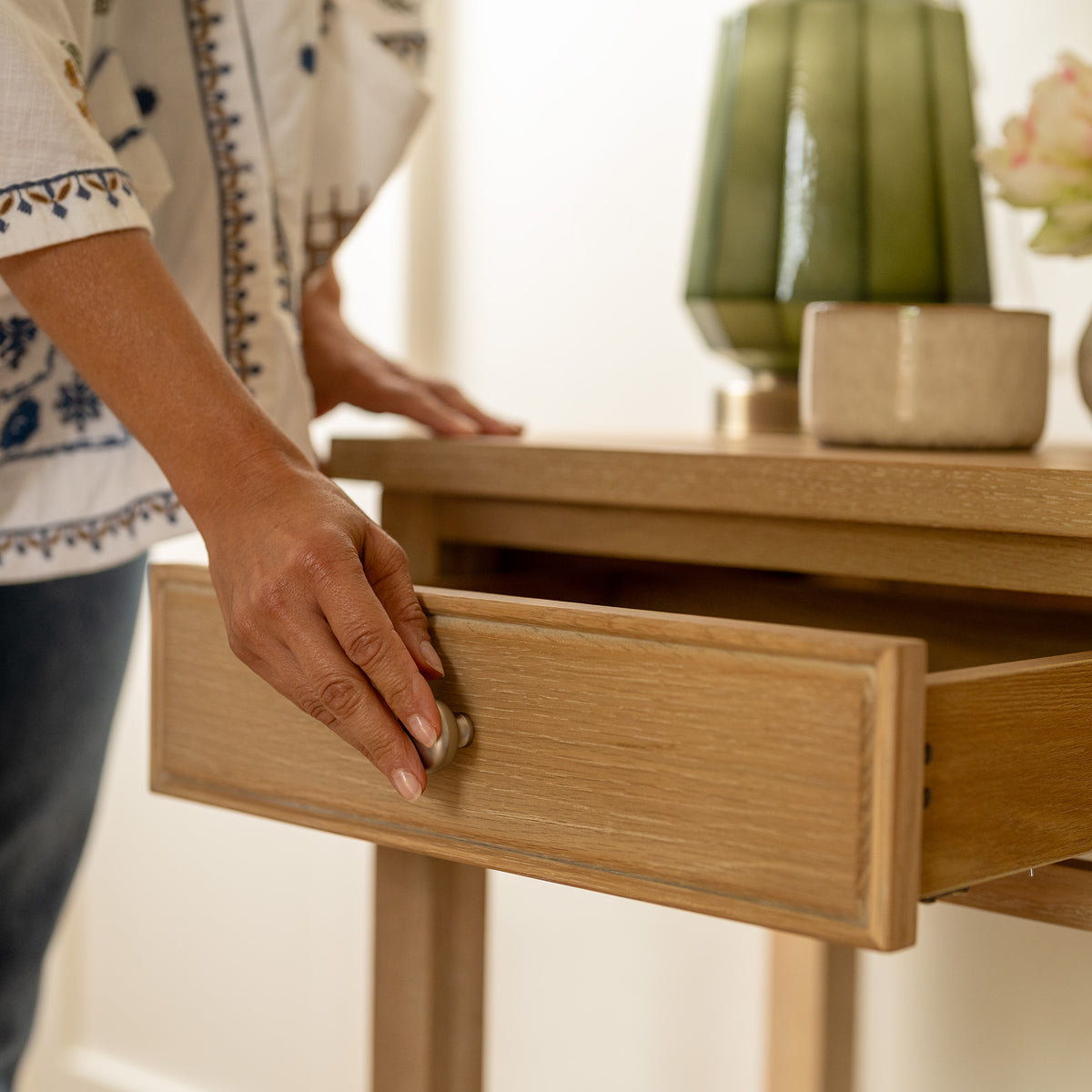 A person opening a drawer of the Amberley 2 Drawers Console Table in Light Oak, with a green lamp and a small beige box placed on top.