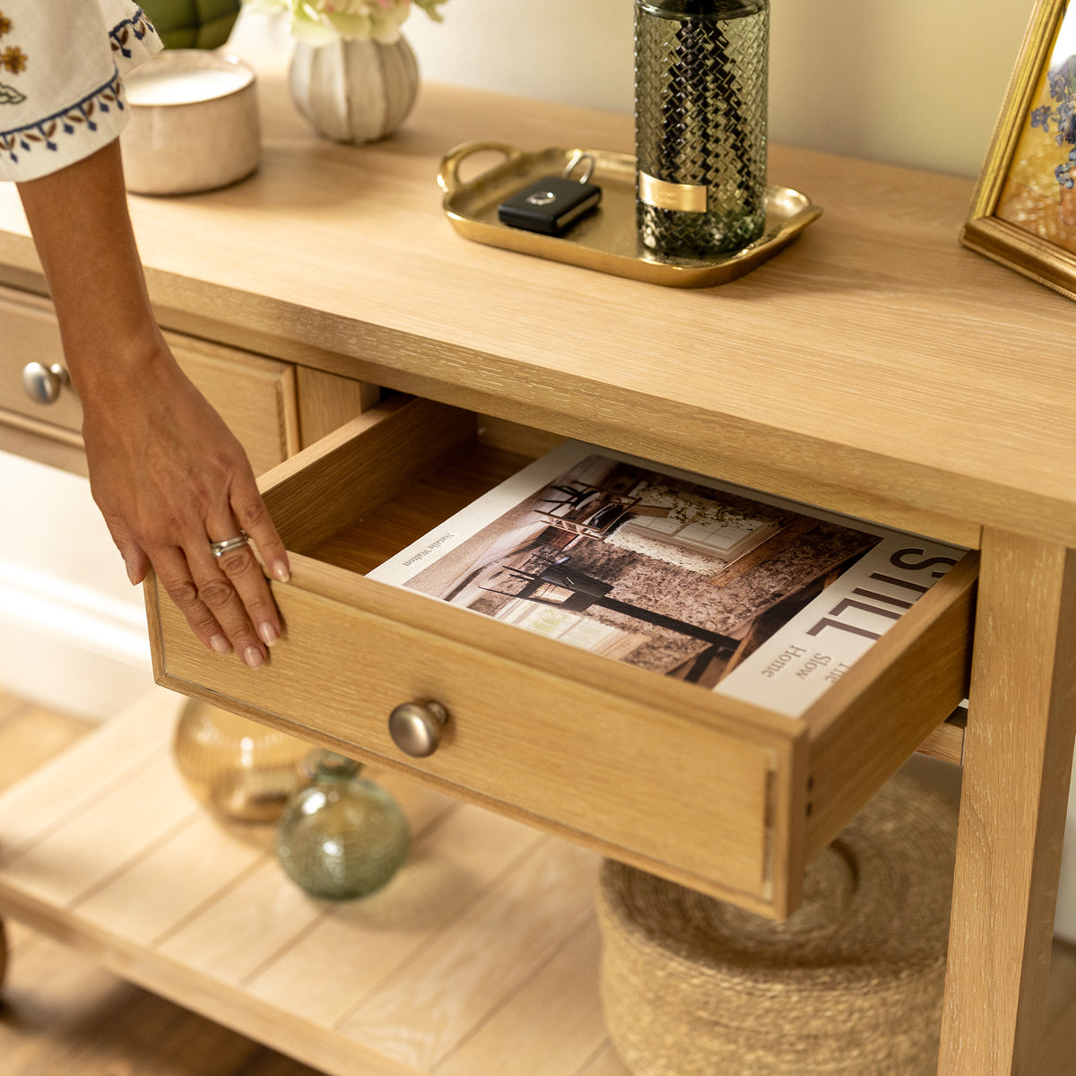 A hand opens a wooden drawer of the Amberley 2 Drawers Console Table in Light Oak, revealing a magazine inside, while decor items and a tray are arranged on top.