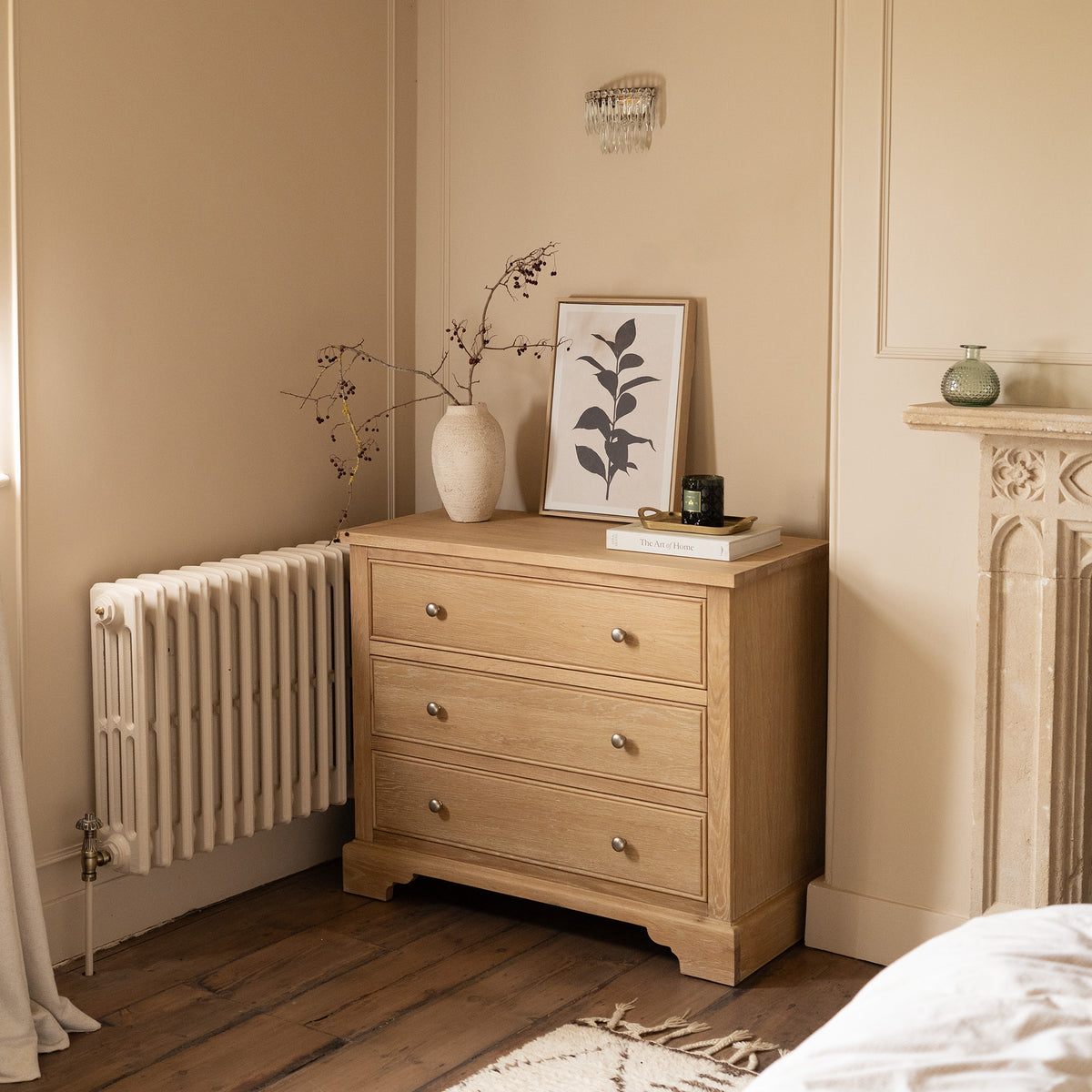 The Amberley 3 Chest of Drawers in Light Oak stands in a cozy, softly lit room, adorned with a vase, artwork, and candle—an ideal choice for stylish bedroom storage.