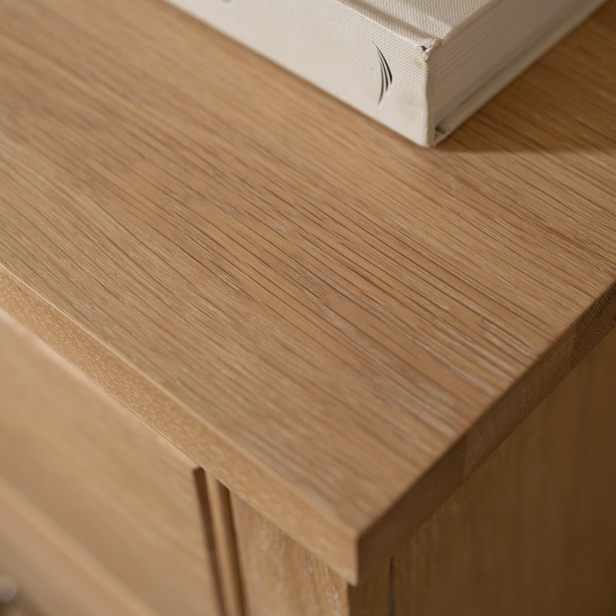 A close-up of the Amberley 3 Chest of Drawers in Light Oak showcases its elegant corner detail with a closed book resting on top—perfect for stylish bedroom storage.