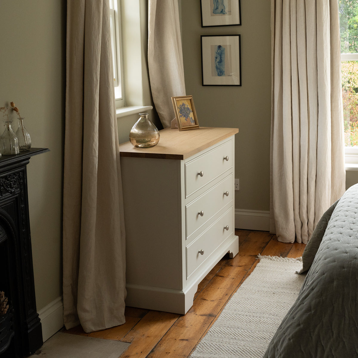 The Amberley 3 Chest of Drawers in Warm White stands elegantly beside a window, its compact form topped with a vase and a framed photo.