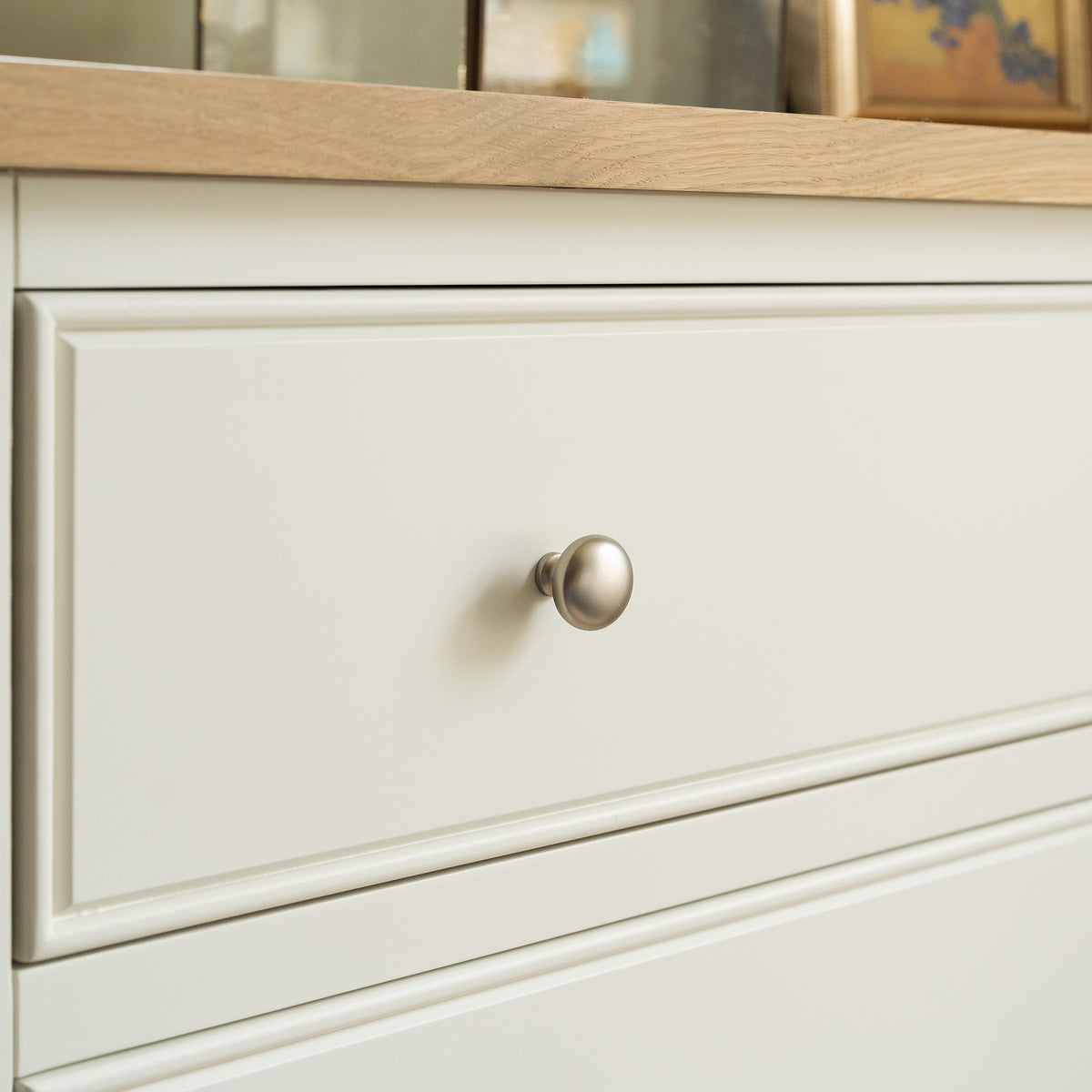 Featuring a warm white finish with silver knobs, this compact chest of drawers is inspired by the classic Amberley 3 Chest of Drawers in Warm White. It showcases a solid oak build with a wooden countertop above and picture frames in the background, blending timeless style with practical storage.