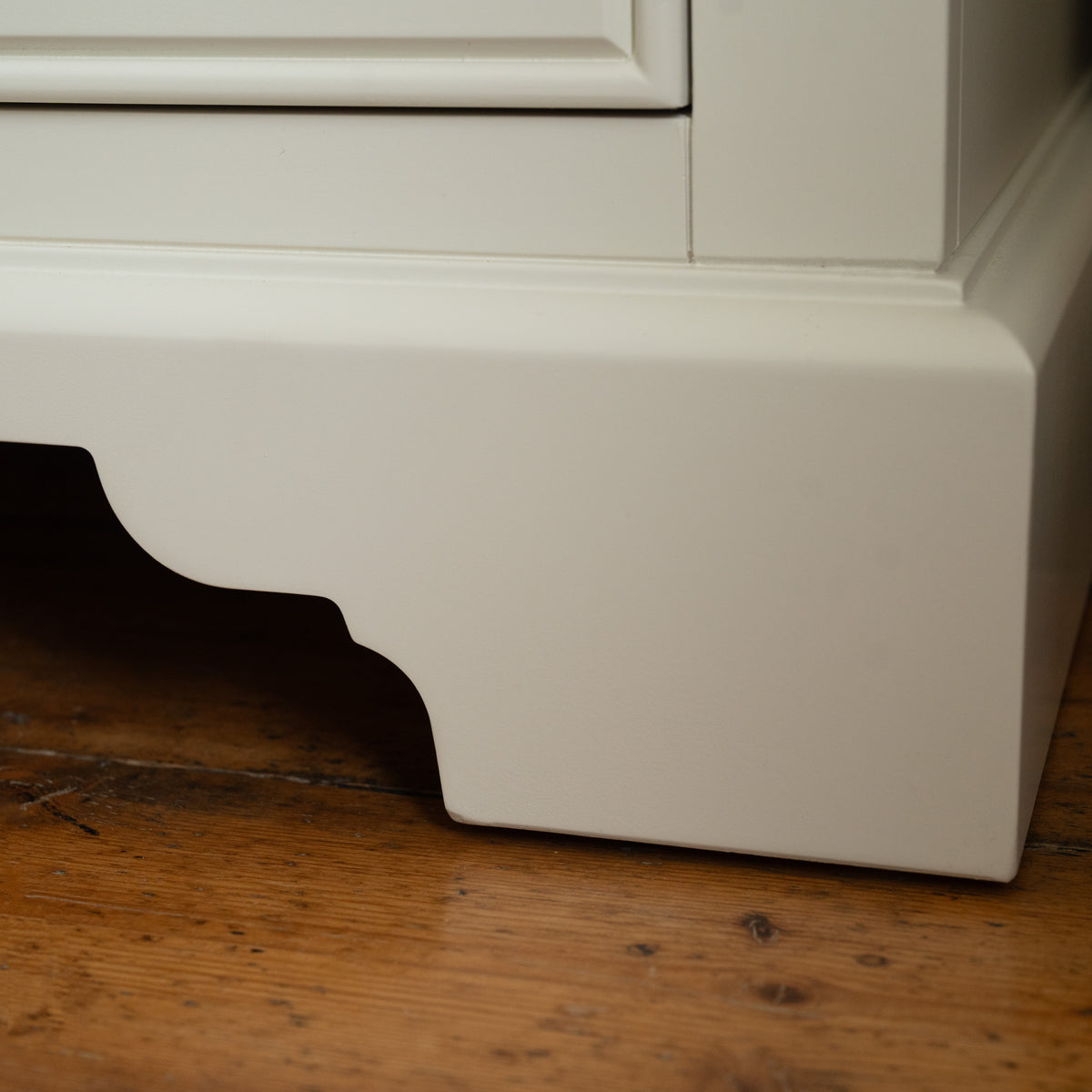Close-up of the bottom corner of a white cabinet on a wooden floor, showcasing the refined design typical of compact storage furniture like the Amberley 3 Chest of Drawers in Warm White.