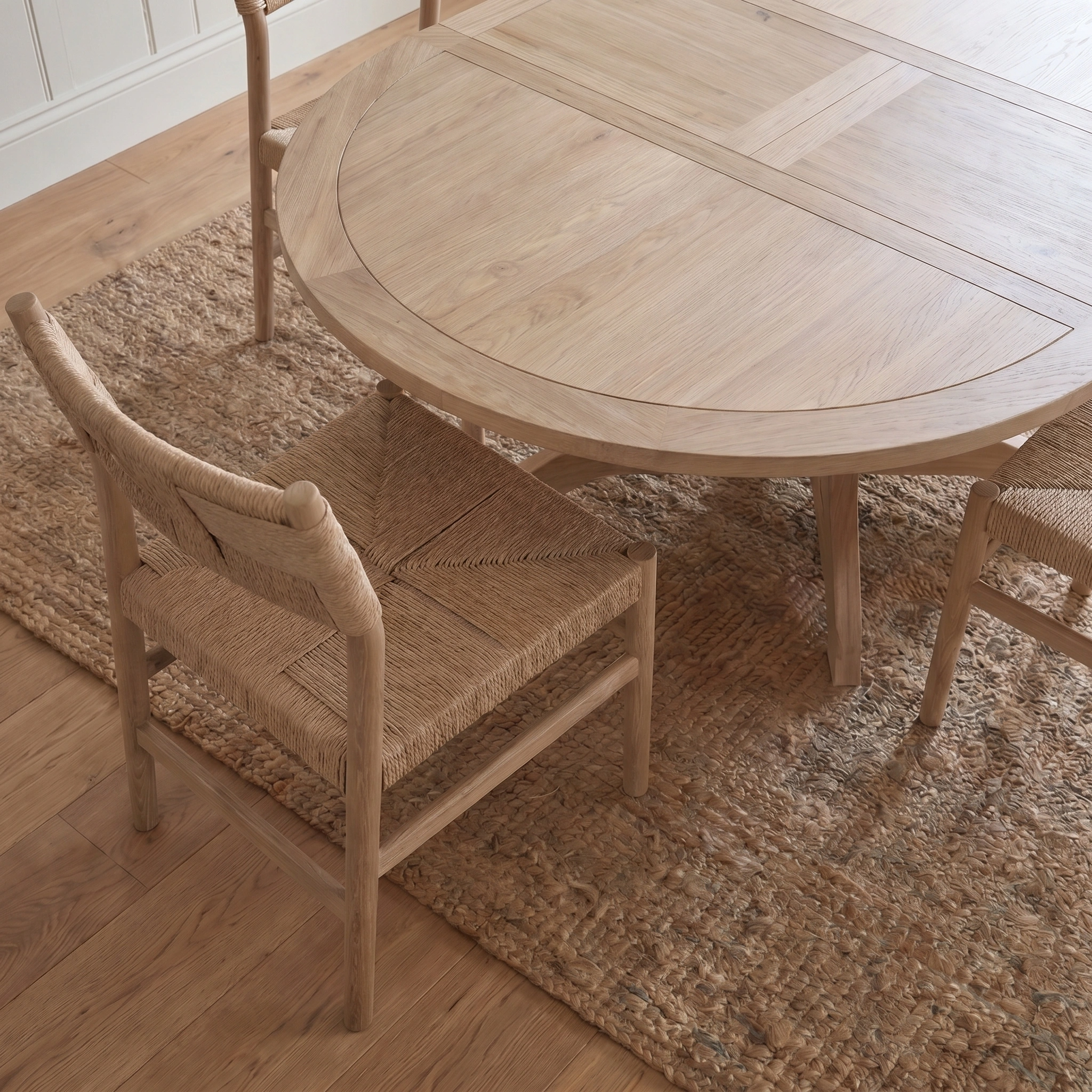 The Amberley 4-6 Seater Round Extending Dining Table in Light Oak with butterfly leaf pairs with woven chairs on a jute rug over hardwood flooring in a bright room.