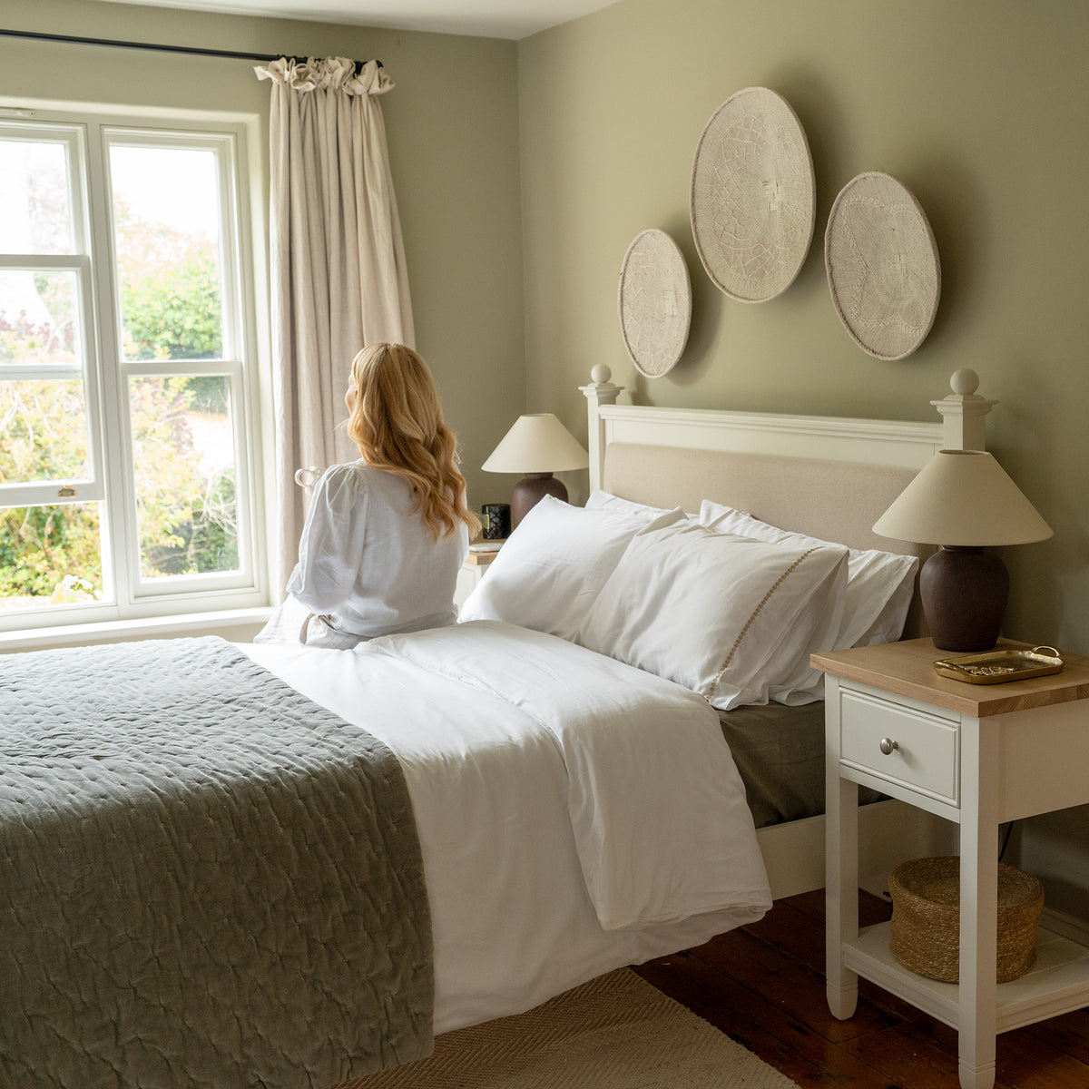 A woman in a white robe sits on a bed near the window in a cozy, light-filled bedroom with neutral decor and an elegant Amberley 5ft Kingsize Bed Frame in Warm White.