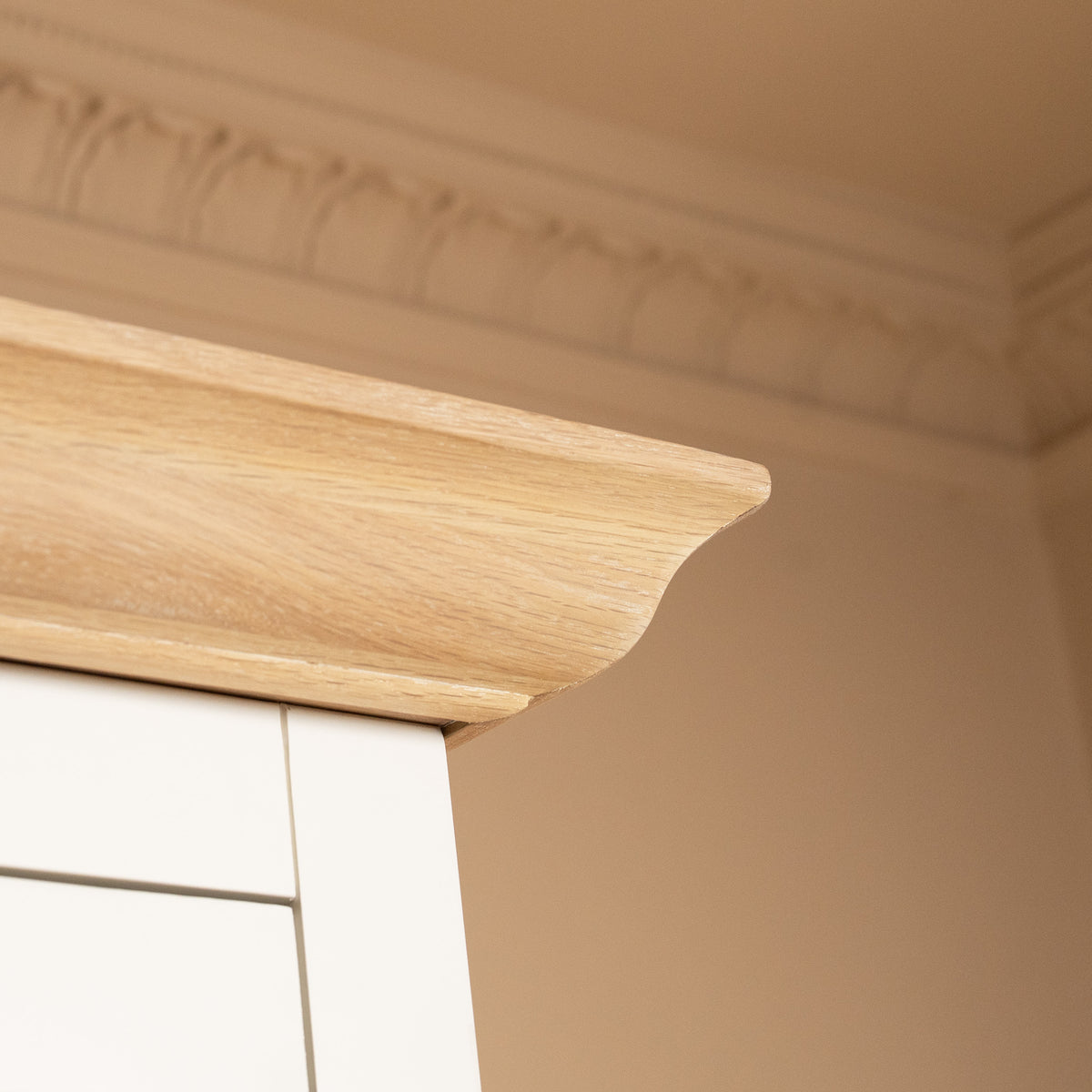 A light wood cabinet corner with crown molding, set against a beige wall with decorative ceiling trim, perfectly complements the Amberley Double Wardrobe in Warm White.