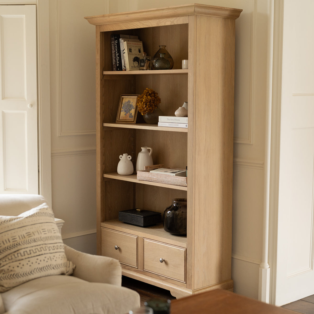 Amberley Large Bookcase in Light Oak, styled with books, vases, framed art, and drawers, stands next to a cream armchair in a cozy room.