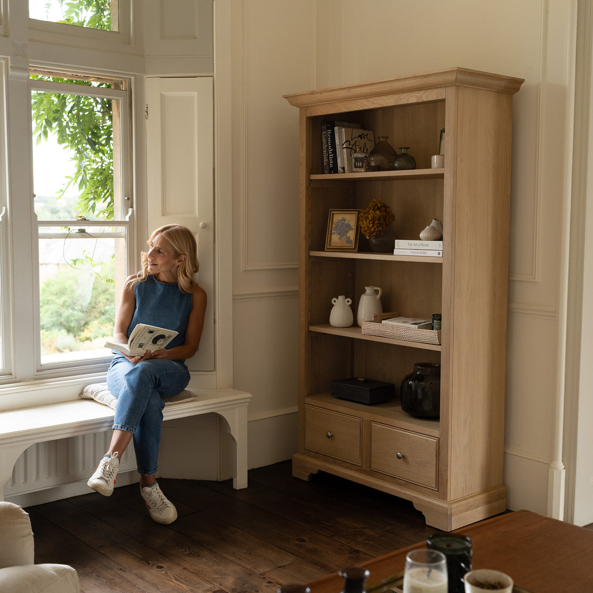 A woman sits by a window reading a book in a cozy room with a modern country vibe, surrounded by charming decor and the Amberley Large Bookcase in Light Oak.