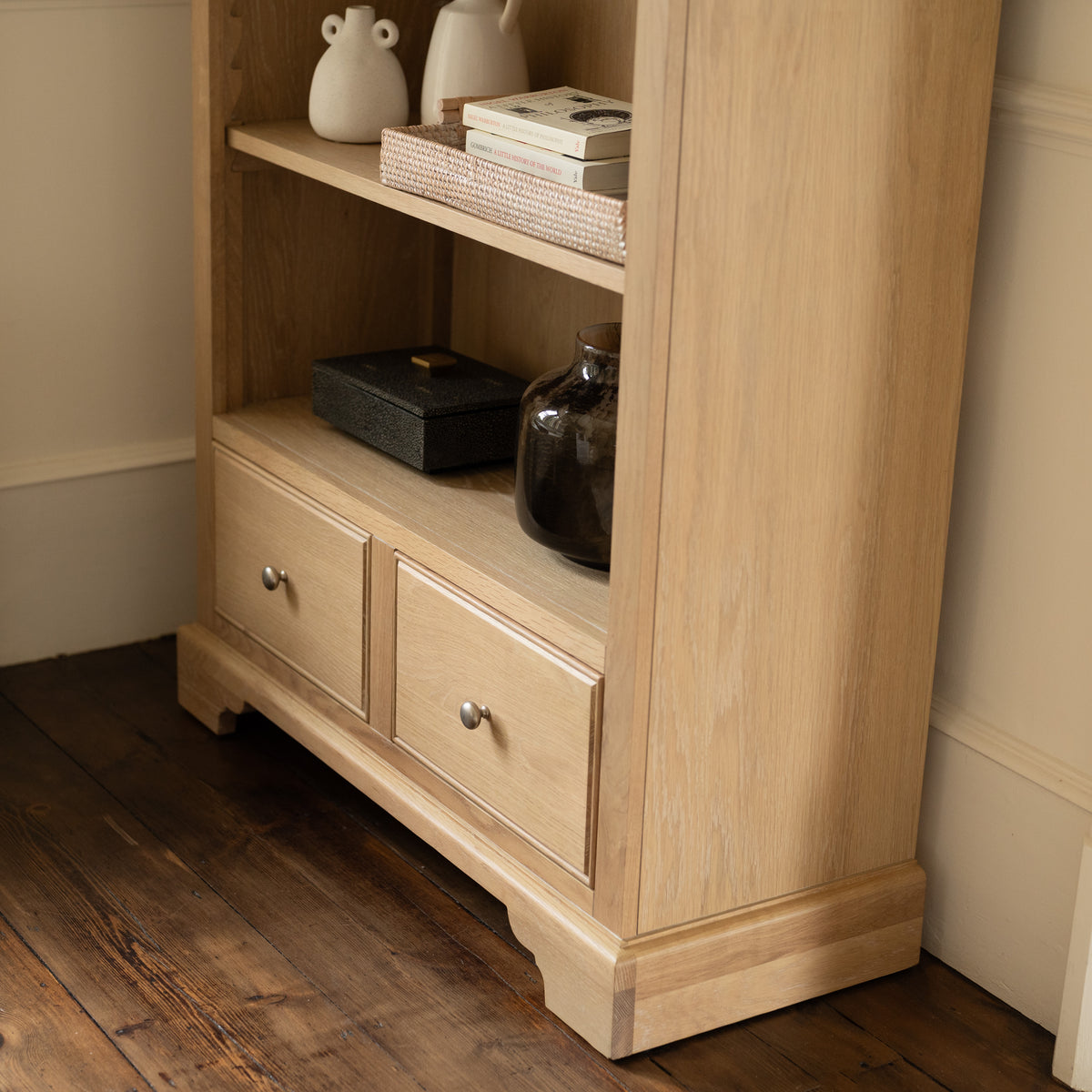 Amberley Large Bookcase in Light Oak featuring shelves and two drawers, styled with books, a vase, and decorative accents on a wooden floor. This bookcase infuses modern country charm into your space with its clean lines and versatile display options.