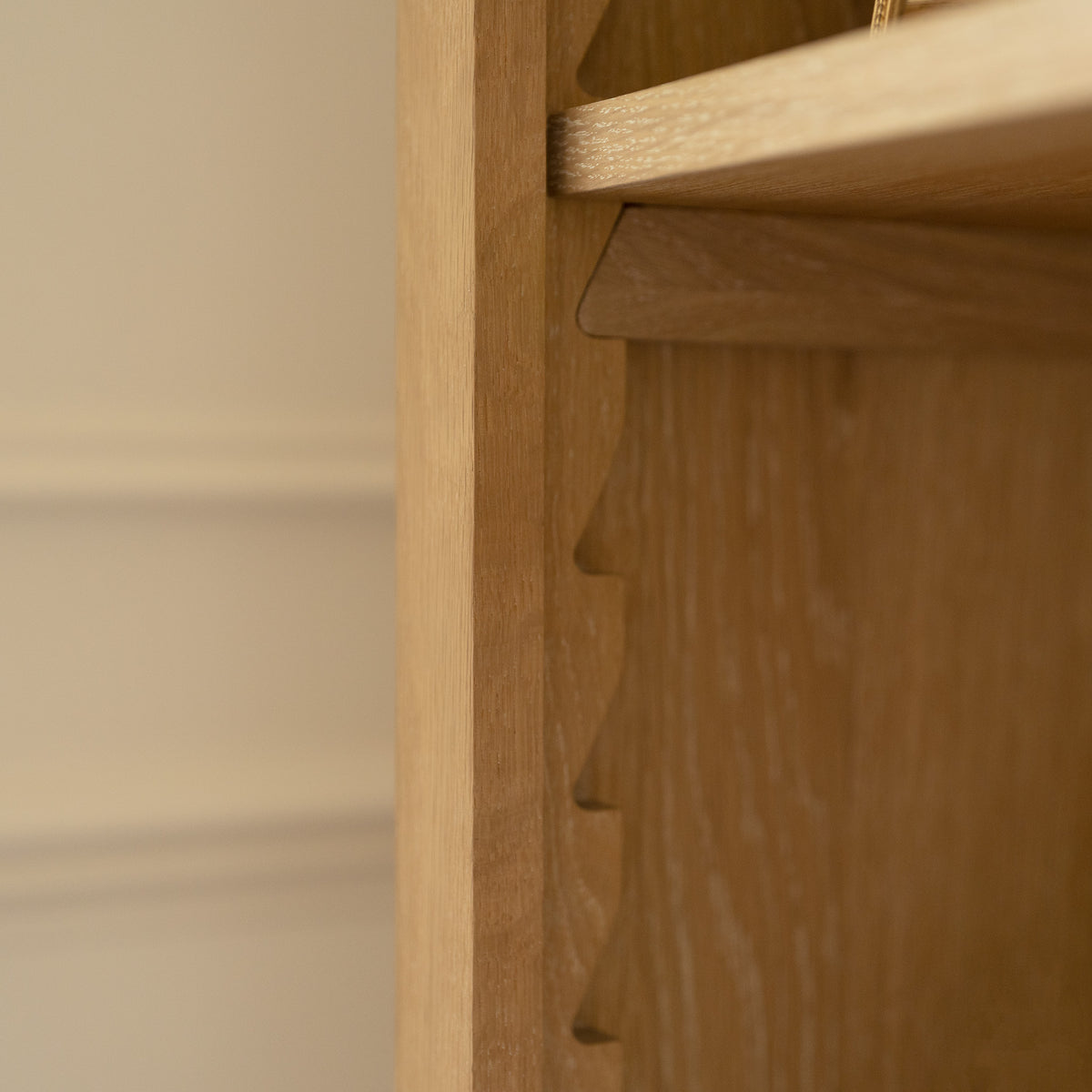 Close-up of a wooden shelf edge with carved grooves, part of the Amberley Large Bookcase in Light Oak, set against a beige wall.