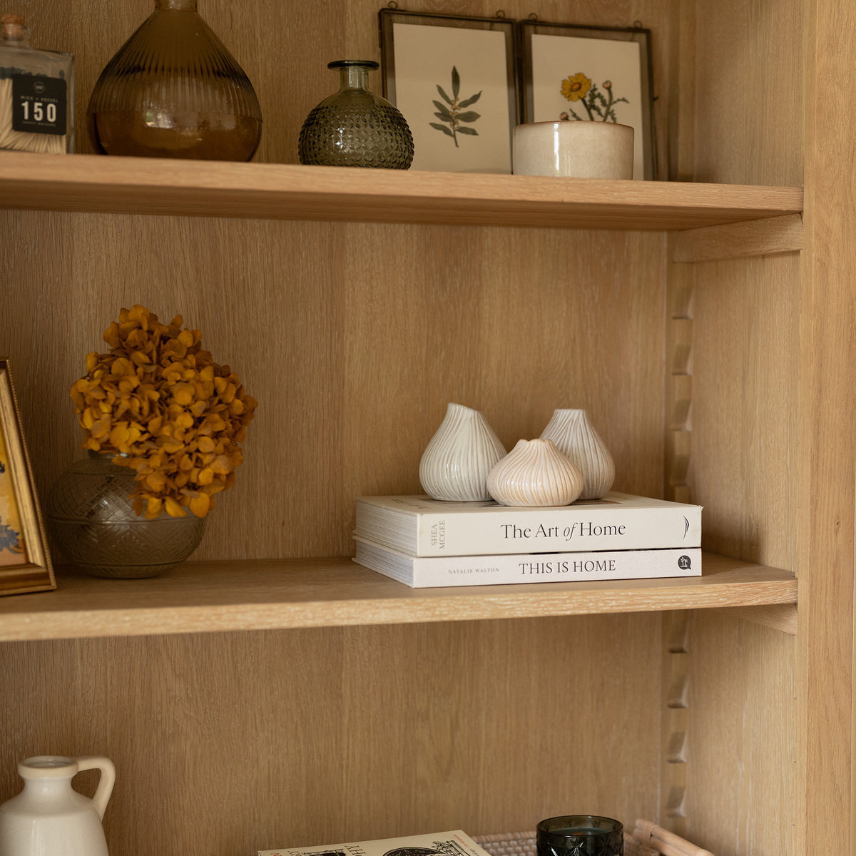 Wooden shelves filled with books, vases, dried flowers, and framed botanical art in a cozy home setting evoke the timeless charm of the Amberley Large Bookcase in Light Oak.