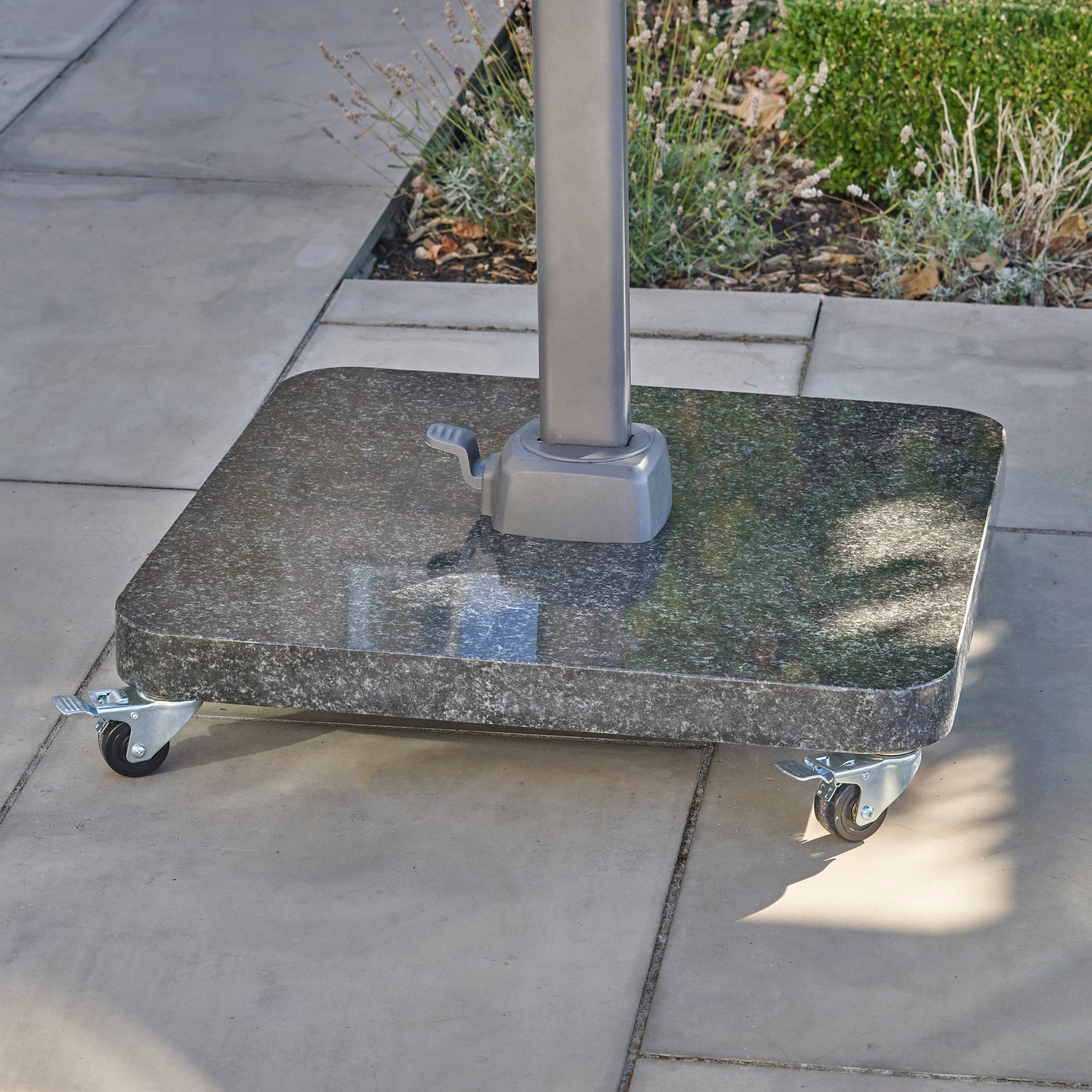 The Ancona 90kg Granite Base with Wheels stands on a tiled outdoor patio near garden plants, providing sturdy support for your umbrella.