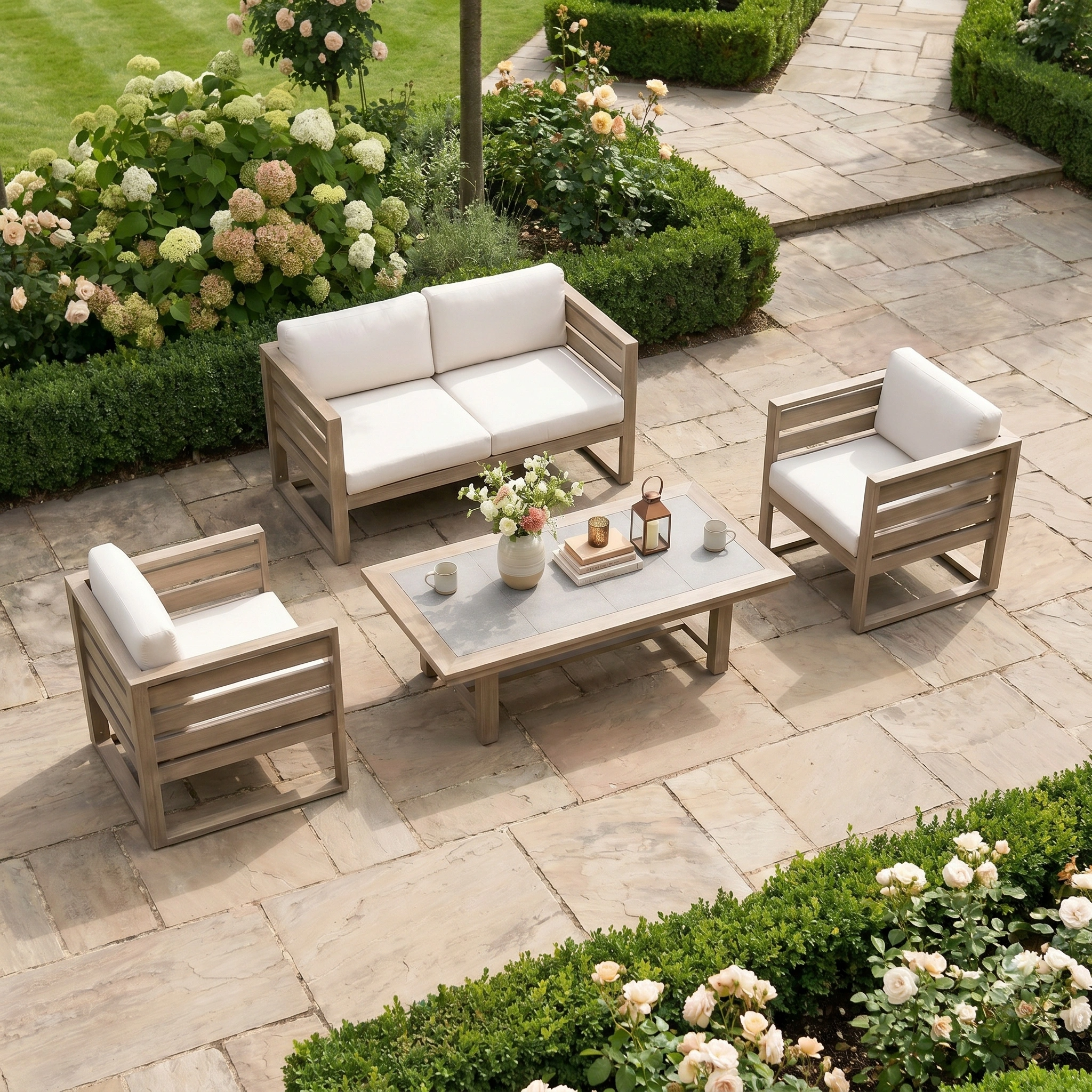The Andora 2 Seat Sofa Set with 2 in 1 Table in Weathered Brown features white cushions and elegant eucalyptus wood, creating an inviting seating area on a stone patio amid lush greenery and flowers.