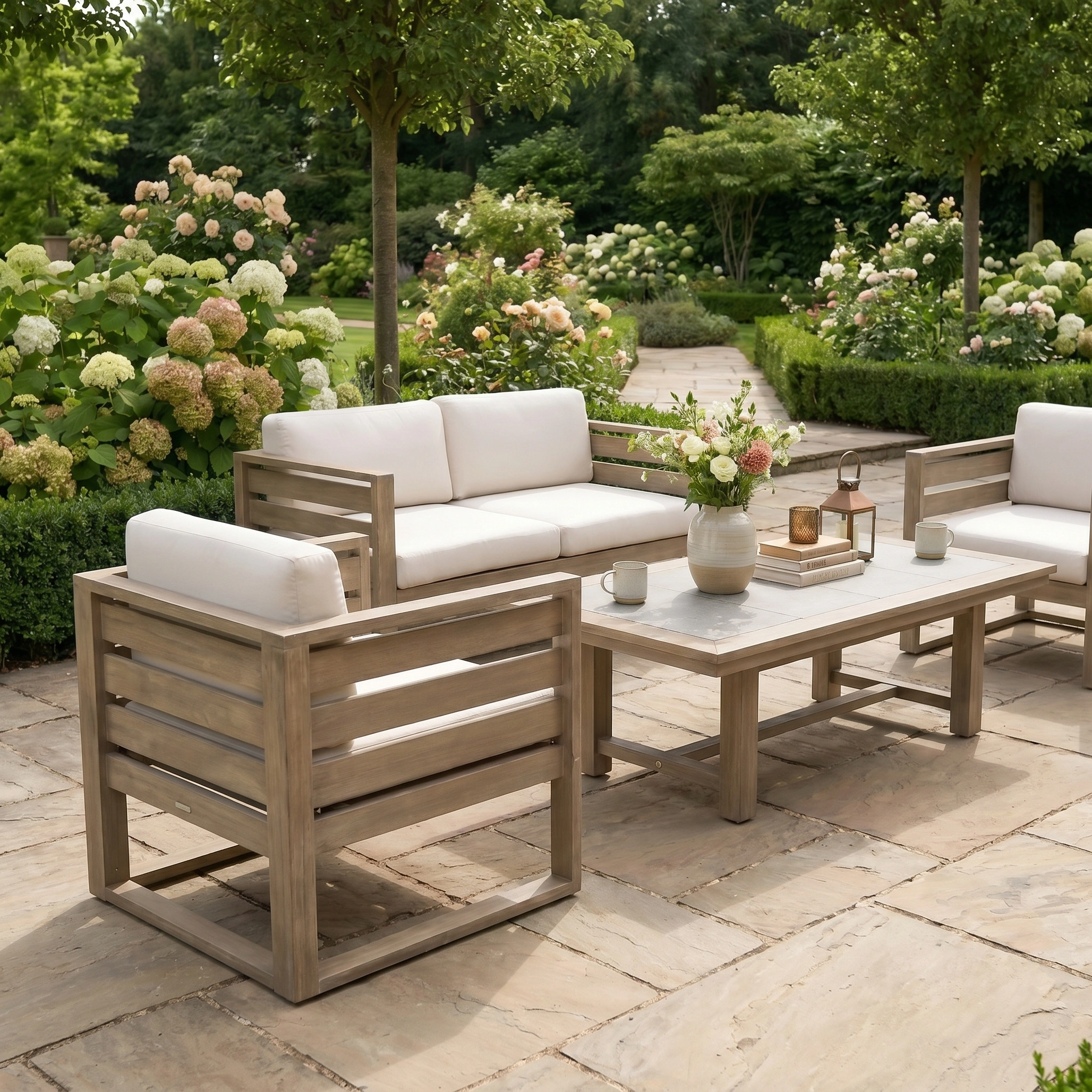 The Andora 2 Seat Sofa Set with 2 in 1 Table in Weathered Brown, paired with white cushions, creates a stylish patio seating area on a stone terrace surrounded by blooming flowers and lush greenery.