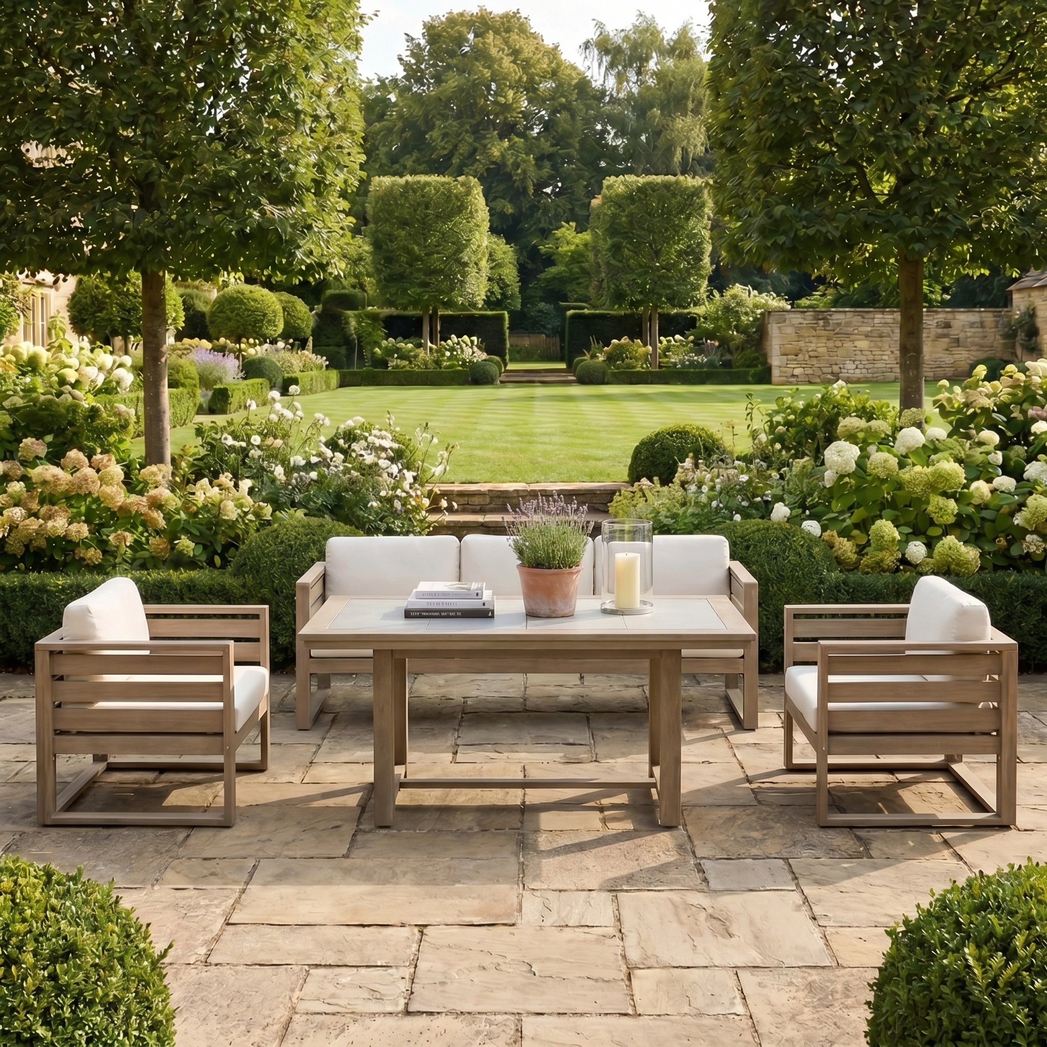 The Andora 3 Seat Sofa Set with 2 in 1 Table in Weathered Brown features white cushions and modular outdoor design, perfect for a landscaped garden surrounded by trees and blooming flowers.