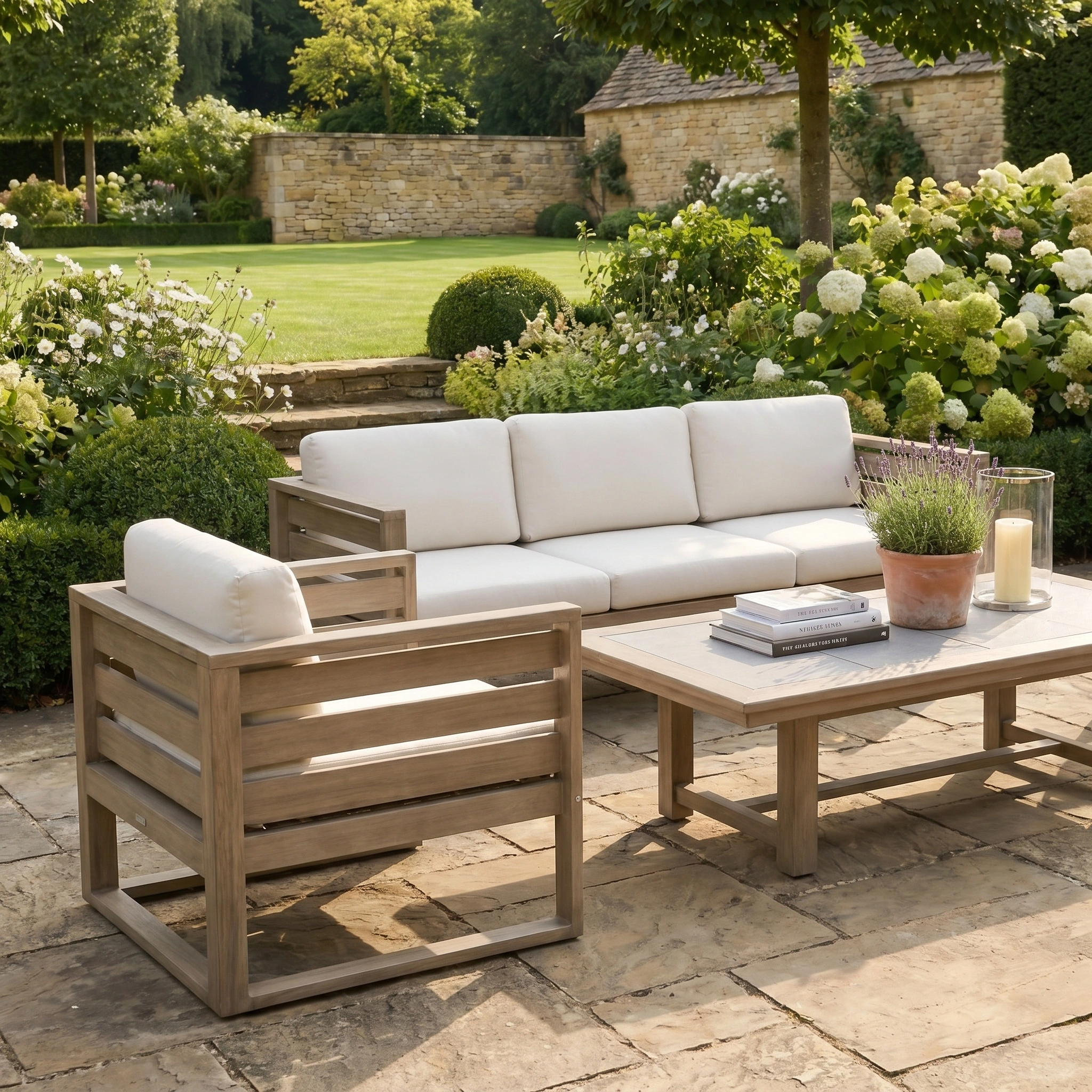 The Andora 3 Seat Sofa Set with 2 in 1 Table in Weathered Brown, featuring white cushions, is arranged on a stone terrace amidst a lush green garden and vibrant flowers.