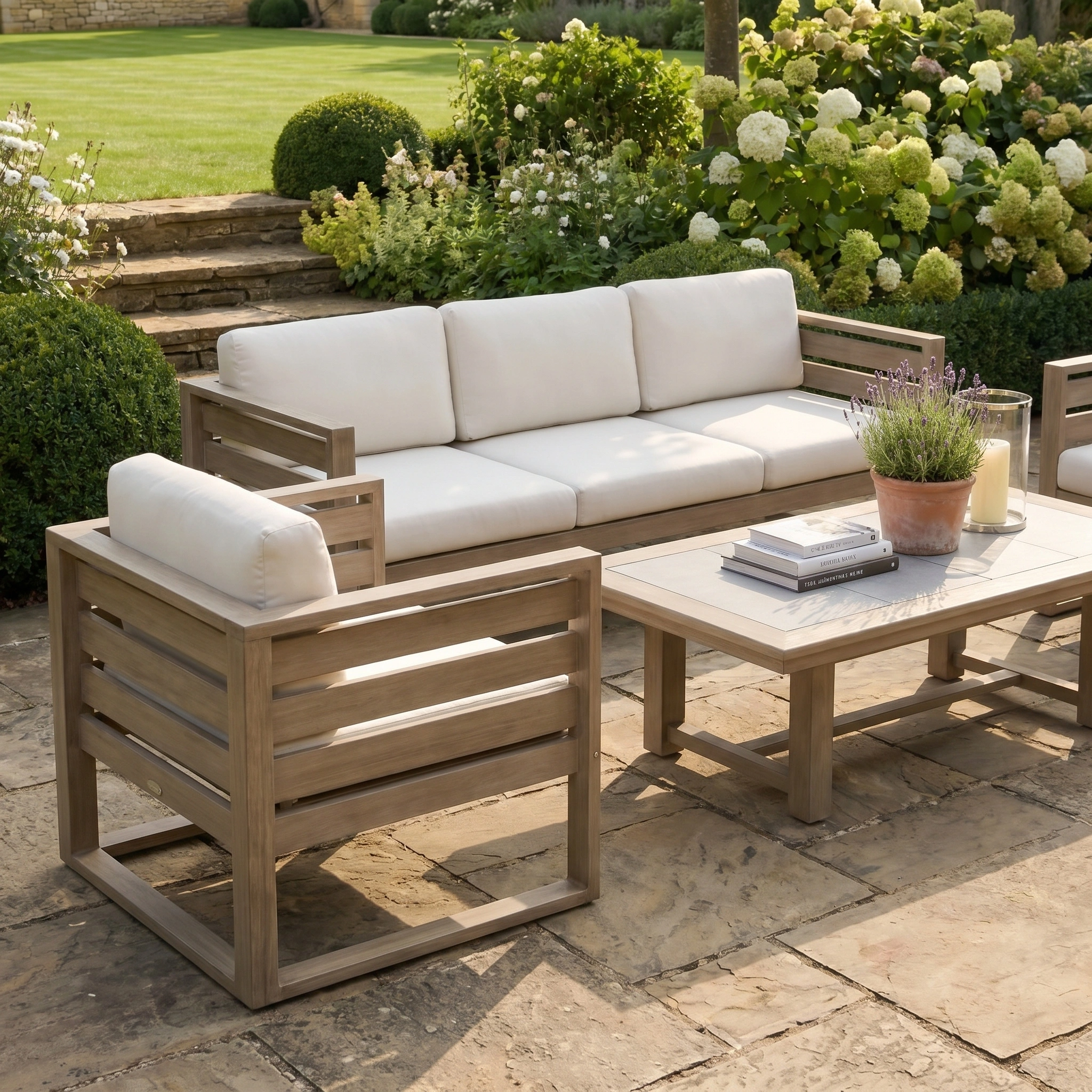 The Andora 3 Seat Sofa Set with 2 in 1 Table in Weathered Brown features modern modular outdoor furniture with white cushions on a stone terrace, surrounded by lush green plants.