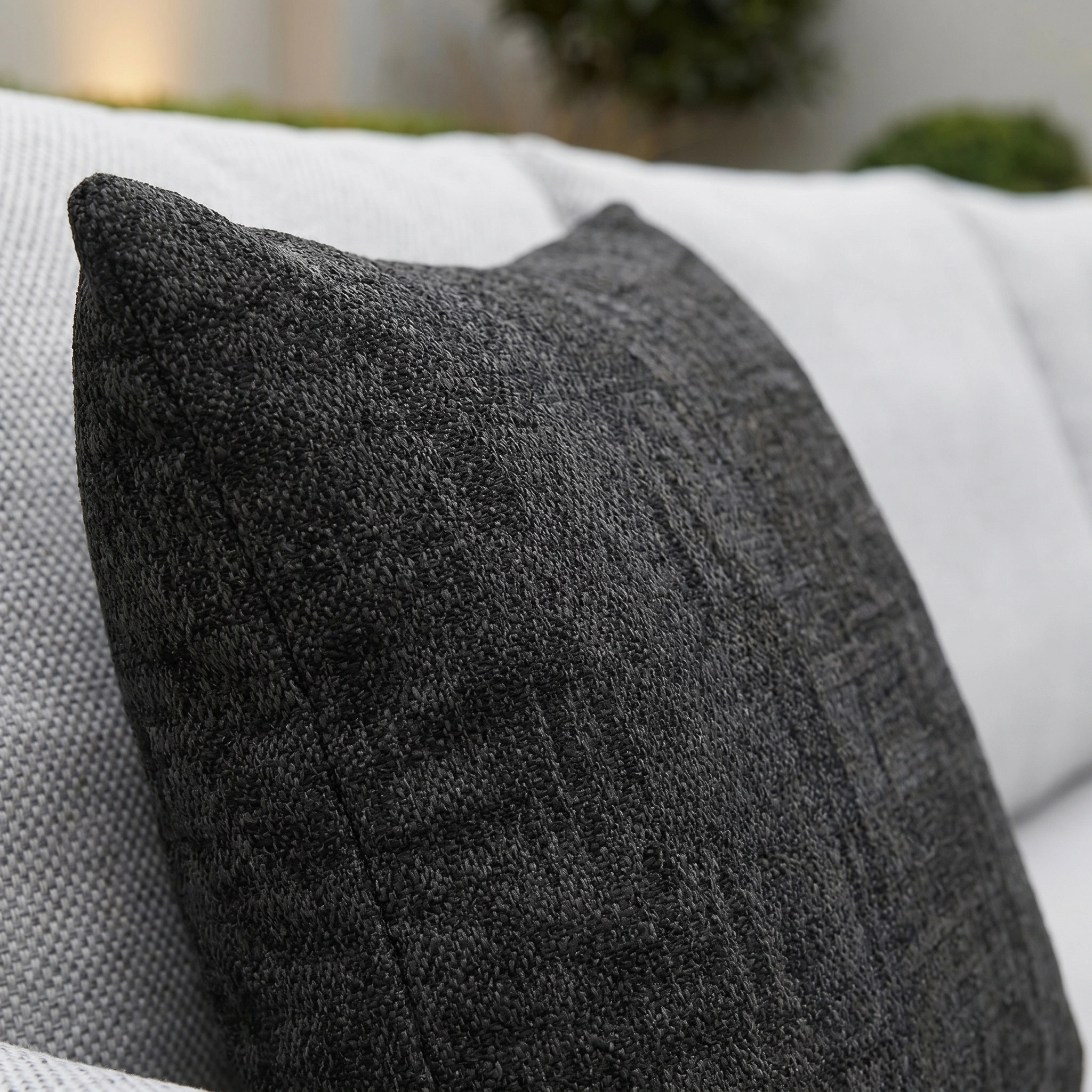The Agora Artisan Grafito Medium Scatter Cushion (45cm x 45cm) features dark gray solution-dyed acrylic on a light gray sofa, with blurred greenery in the background—ideal as an outdoor scatter or stylish garden cushion.