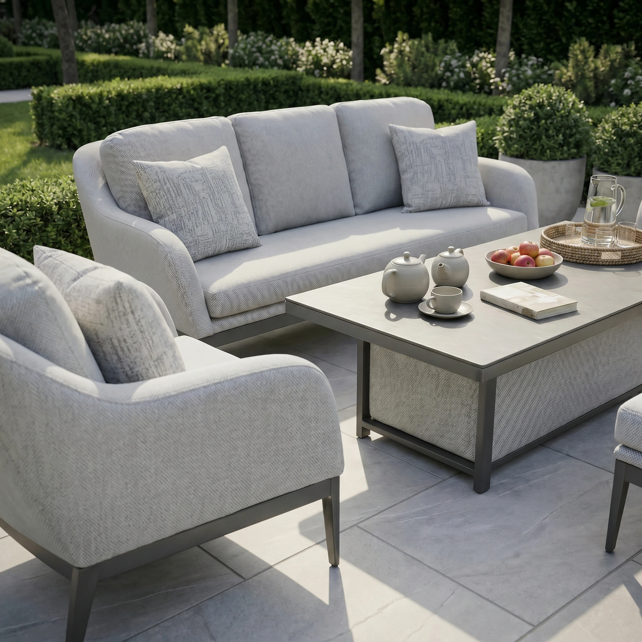 Modern outdoor patio set with light gray sofas featuring Agora Artisan Perla Medium Scatter Cushions (45cm x 45cm), a coffee table, tea set, fruit, and a lush green garden background.