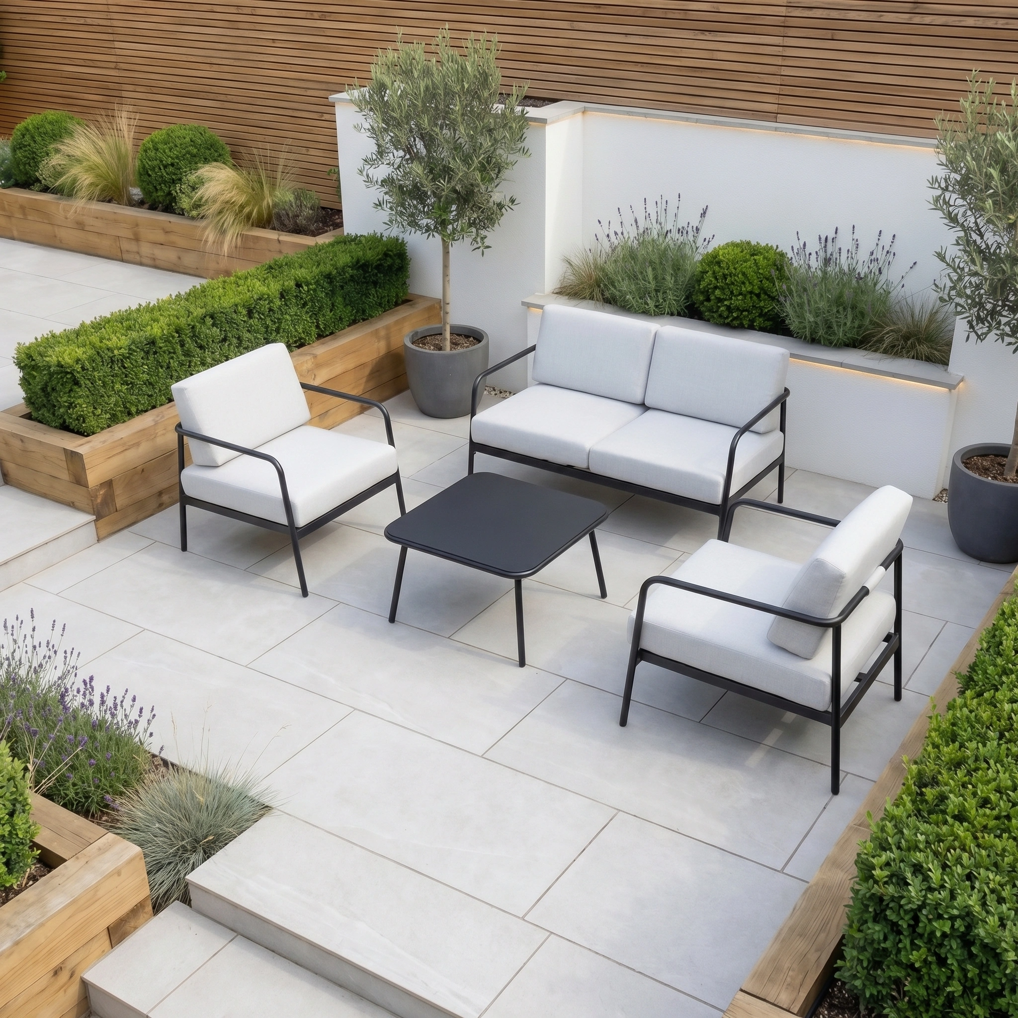 Modern outdoor patio with the Aspen 2 Seat Sofa and Coffee Table in Light Grey, featuring white cushions, a loveseat, chairs, potted plants, and landscaped greenery.