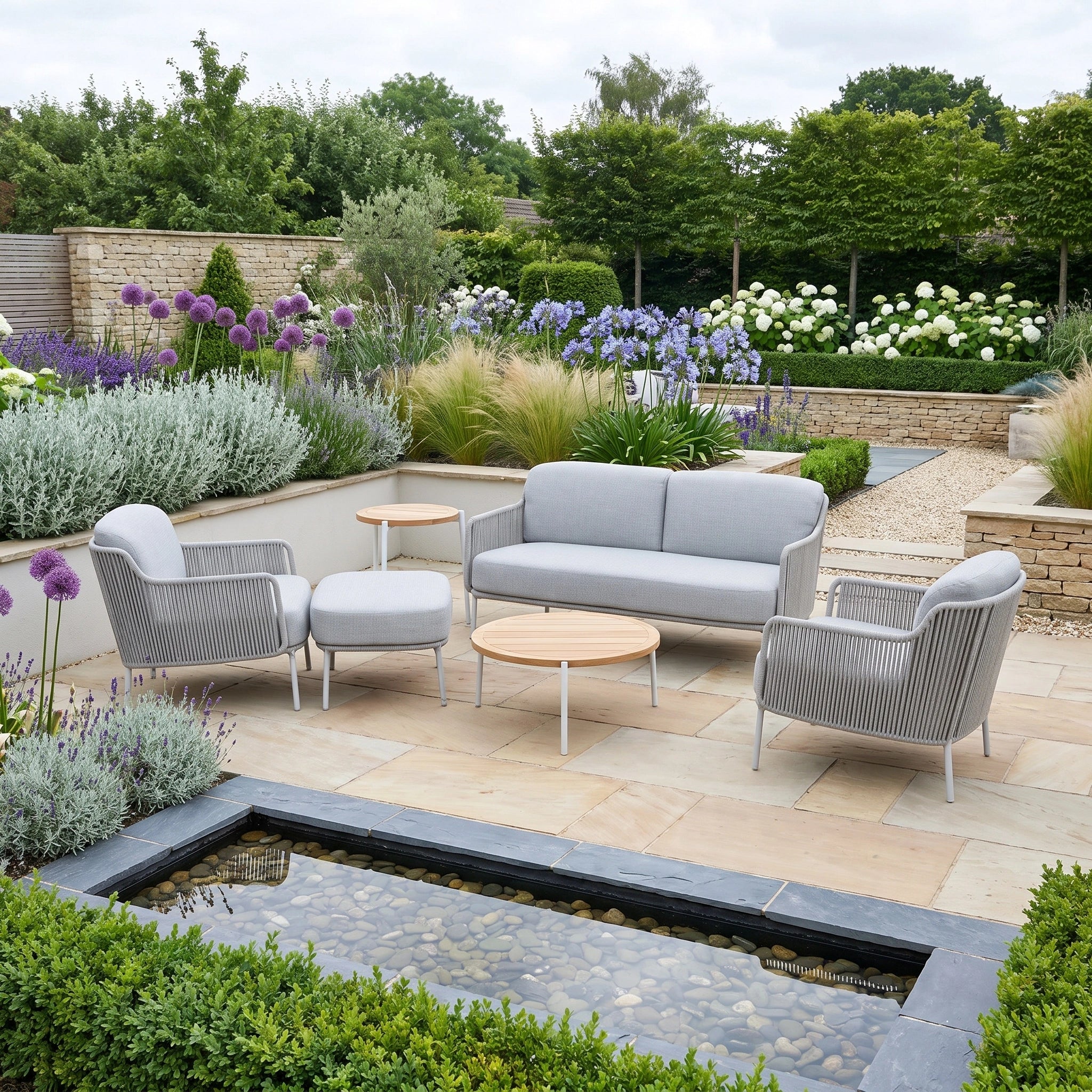 The Bernini 2 Seat Sofa Set with Teak Tables and Footstool in Light Grey arranged on a stone patio, surrounded by lush garden plants and a small pond.