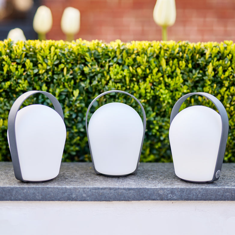 A set of three Bob Small Table Lights in Charcoal, featuring modern, oval-shaped designs with handles, adorn a stone ledge in front of green bushes, serving as elegant garden lamps. These rechargeable and weather-resistant lights are perfect for outdoor settings.