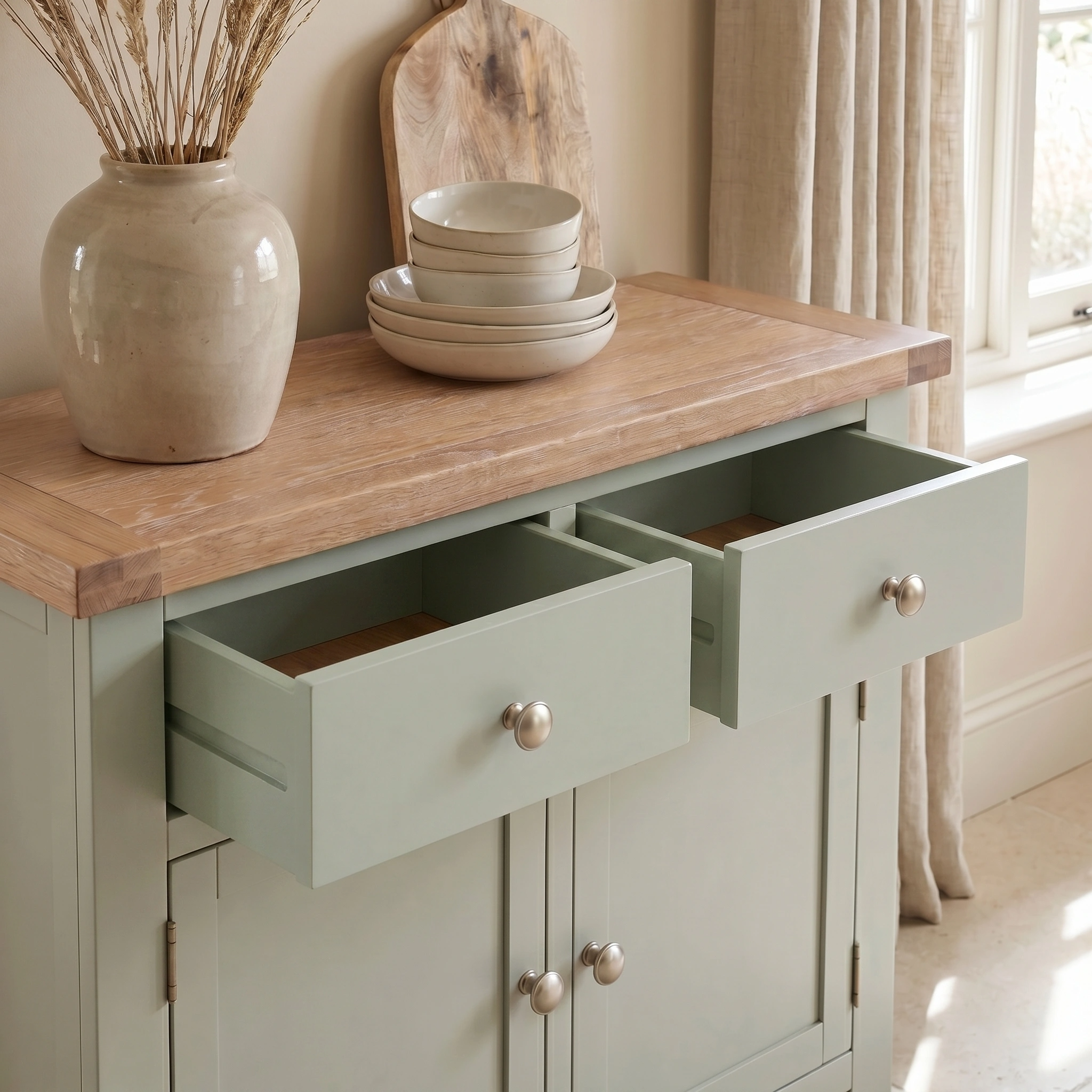 The Burford 2 Door 2 Drawers Sideboard in Sage Green, with open drawers displaying dishes and a vase, is placed on a wooden countertop beside a window.