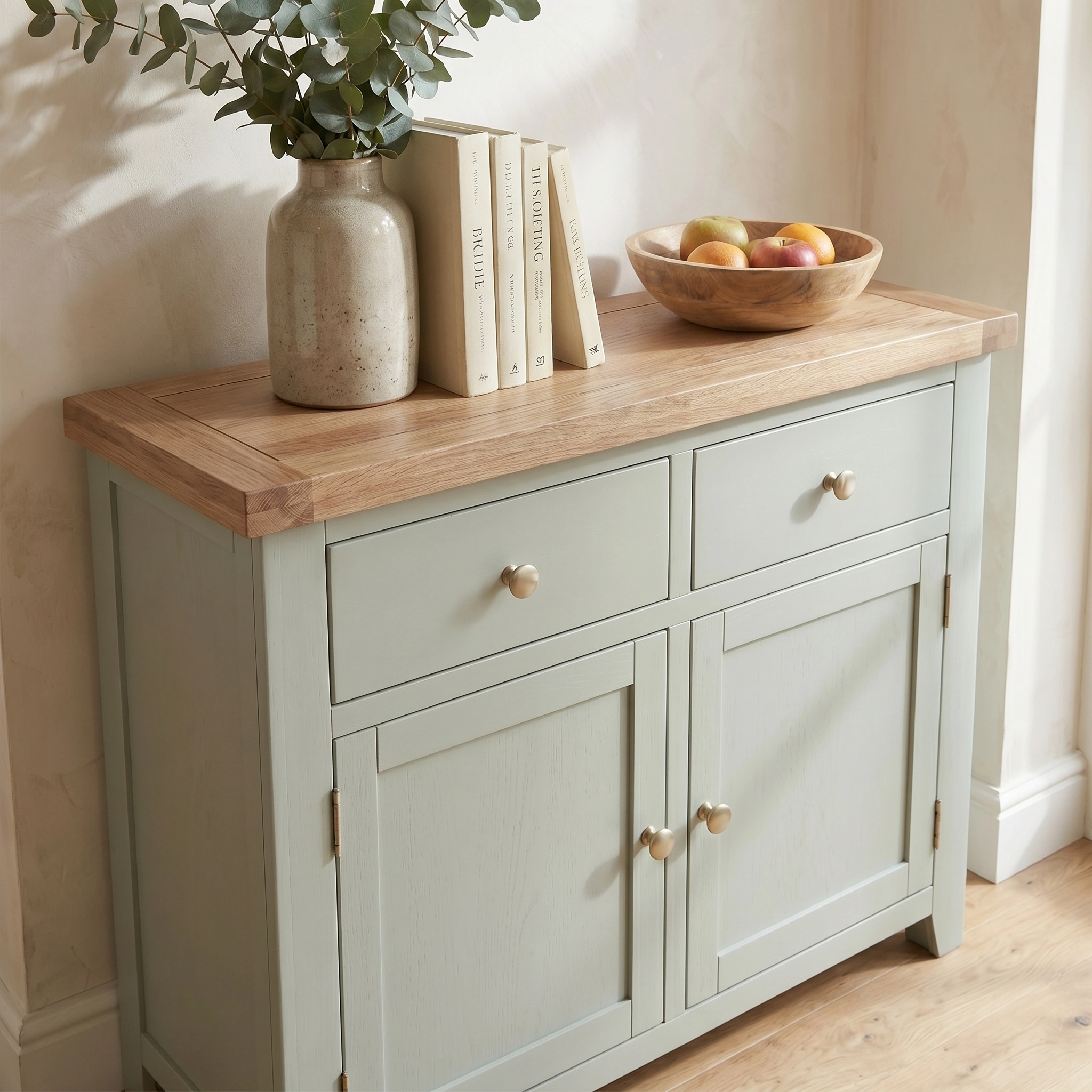 The Burford 2 Door 2 Drawers Sideboard in Sage Green with a wooden top, decorated with books, a vase of leaves, and a fruit bowl, creates a fresh and inviting display.