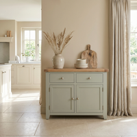 Light-filled kitchen featuring the Burford 2 Door 2 Drawers Sideboard in Sage Green, styled with a ceramic vase, plates, and cutting boards near beige curtains.