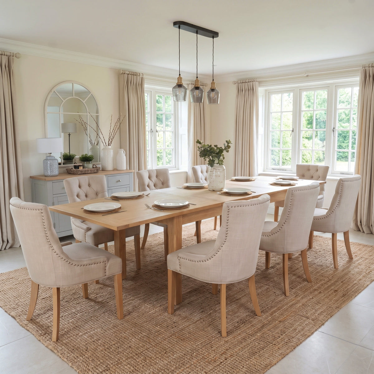 Bright dining room featuring the Burford 8 Seat Extending Dining Set with Regent Chairs in Natural Oak and Ivory, complete with an extendable dining table and eight upholstered chairs. Large windows let in natural light, highlighting the beauty of this natural oak and ivory dining set.