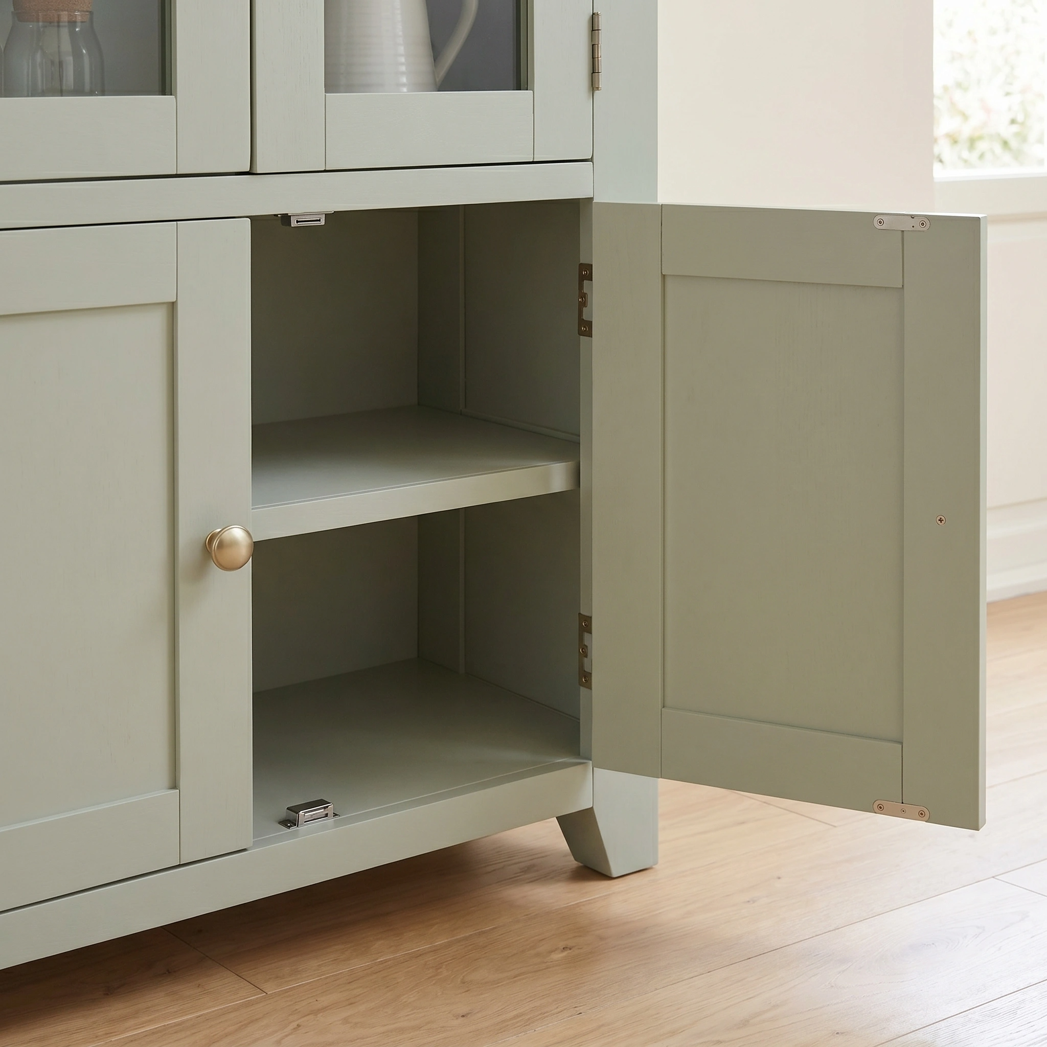 The Burford Display Cabinet in Sage Green, featuring a classic Shaker design, stands open to reveal two empty interior shelves behind its lower door.