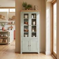 A Burford Display Cabinet in Sage Green with glass doors displays dishes and bowls, placed by a sunlit window in a modern kitchen.