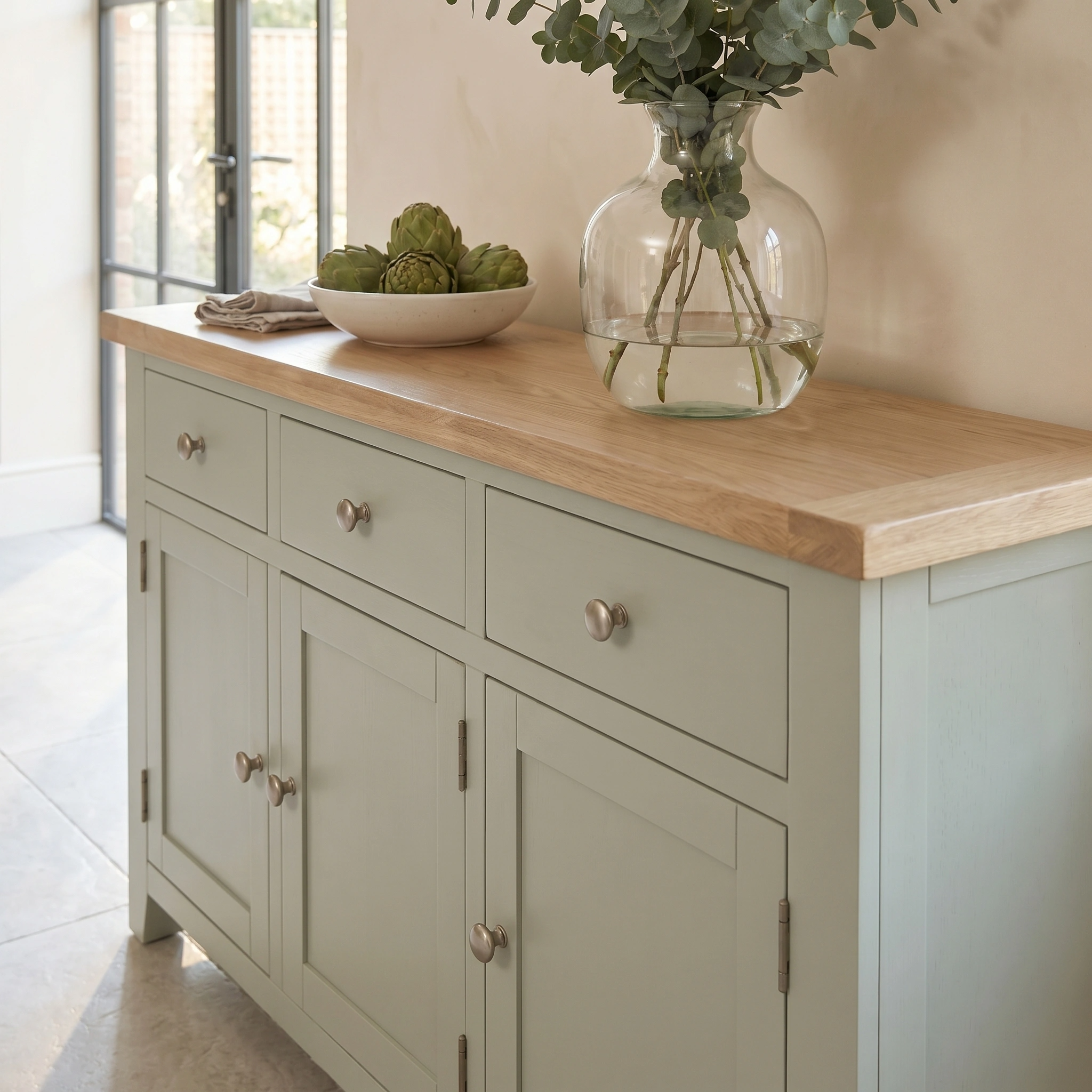 The Burford Large 3 Door 3 Drawers Sideboard in Sage Green features an oak top, decorated with a glass vase of greenery and a bowl of artichokes.
