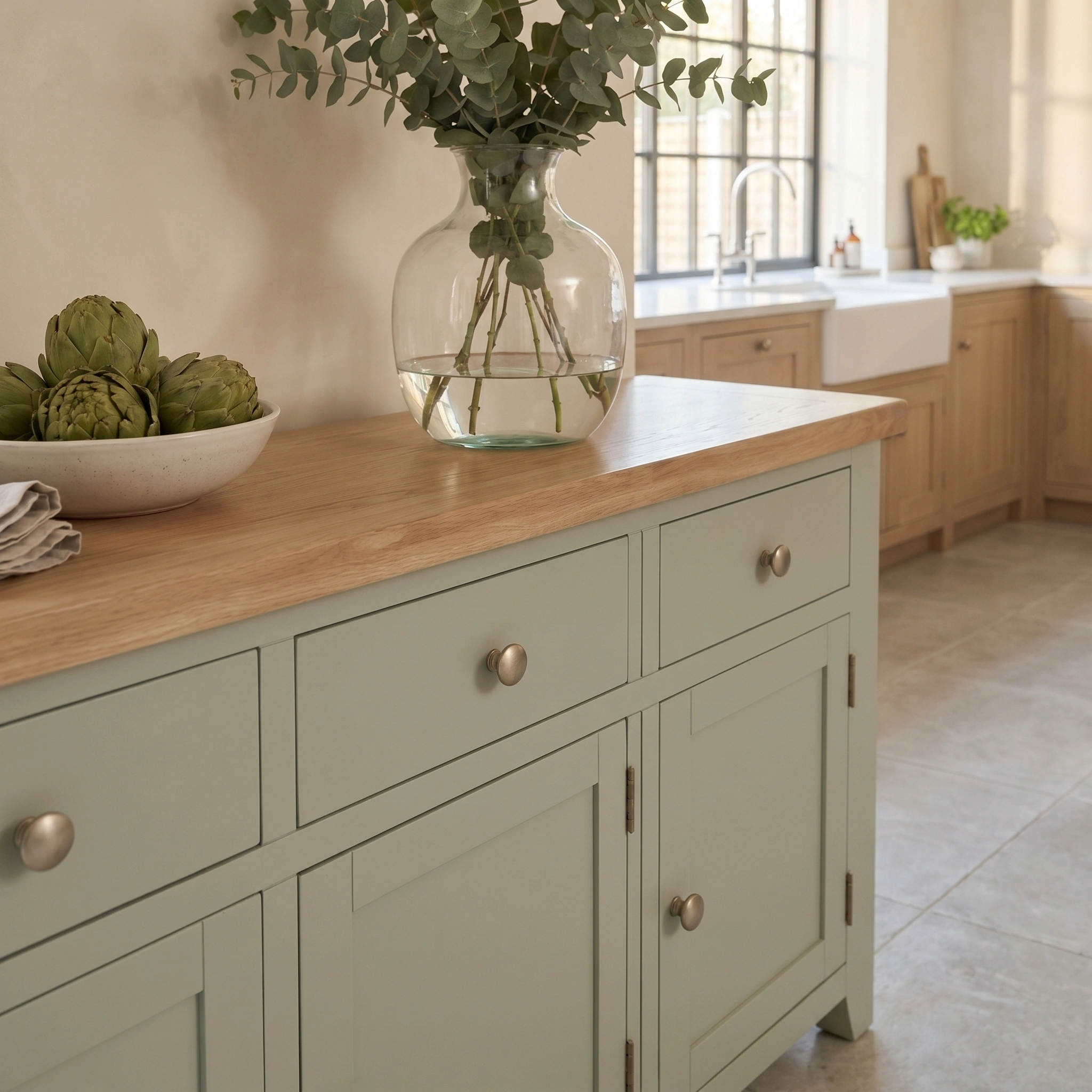 The Burford Large 3 Door 3 Drawers Sideboard in Sage Green is styled with a vase of greenery and a bowl of artichokes, creating a fresh look in a bright kitchen.