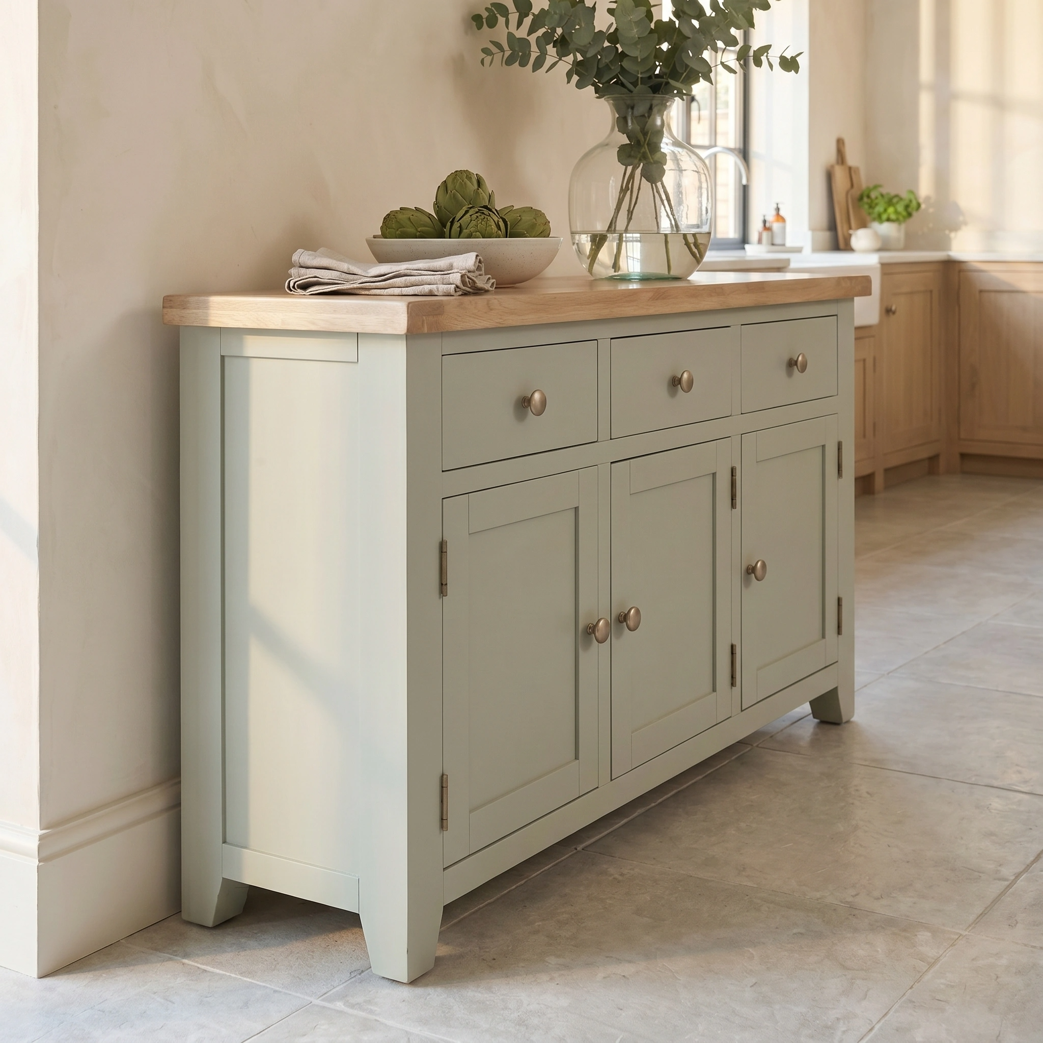 The Burford Large 3 Door 3 Drawers Sideboard in Sage Green features an oak top, drawers, and cabinets, styled with a vase and folded linens.