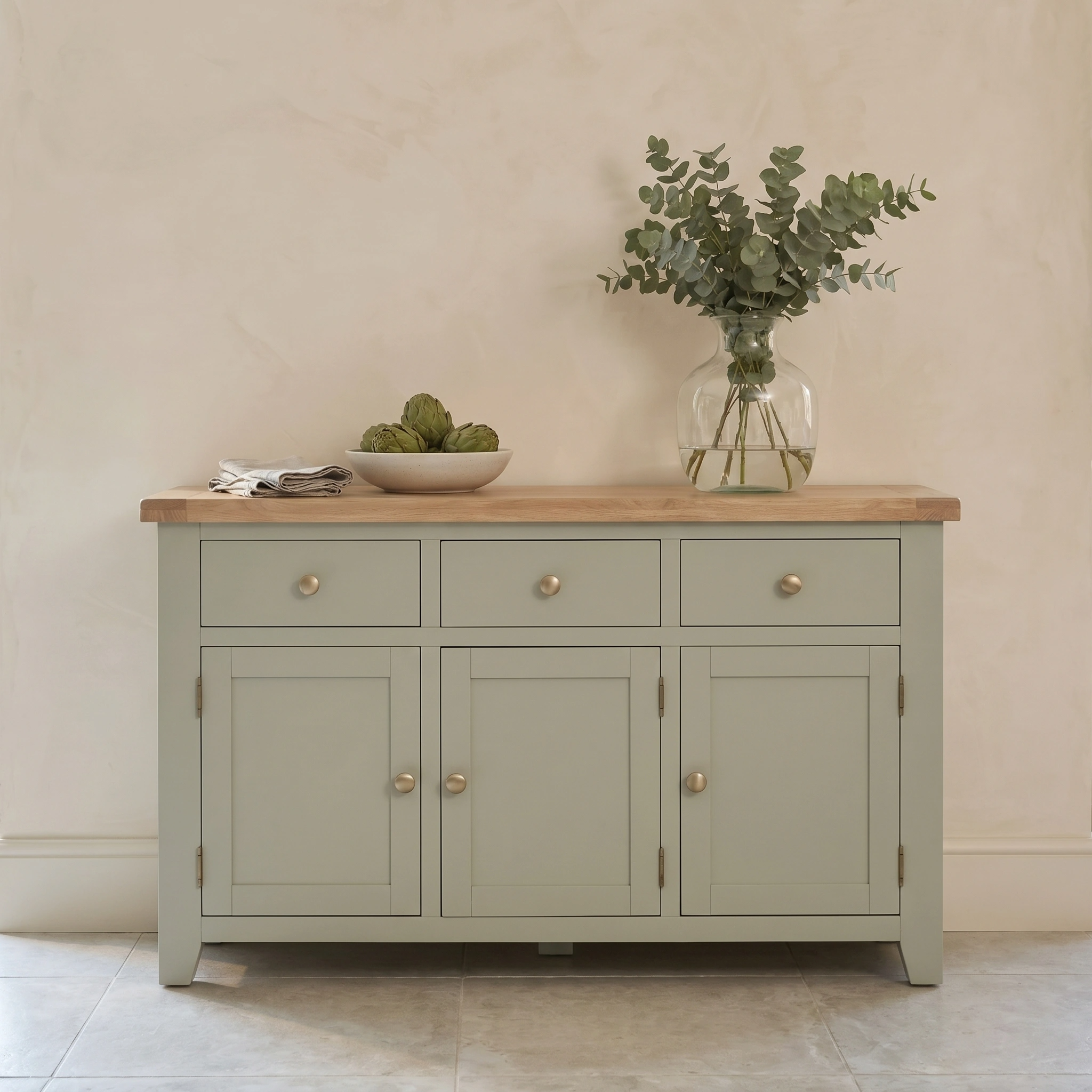 The Burford Large 3 Door 3 Drawers Sideboard in Sage Green features a wooden top, styled with a vase of greenery and a bowl of artichokes.