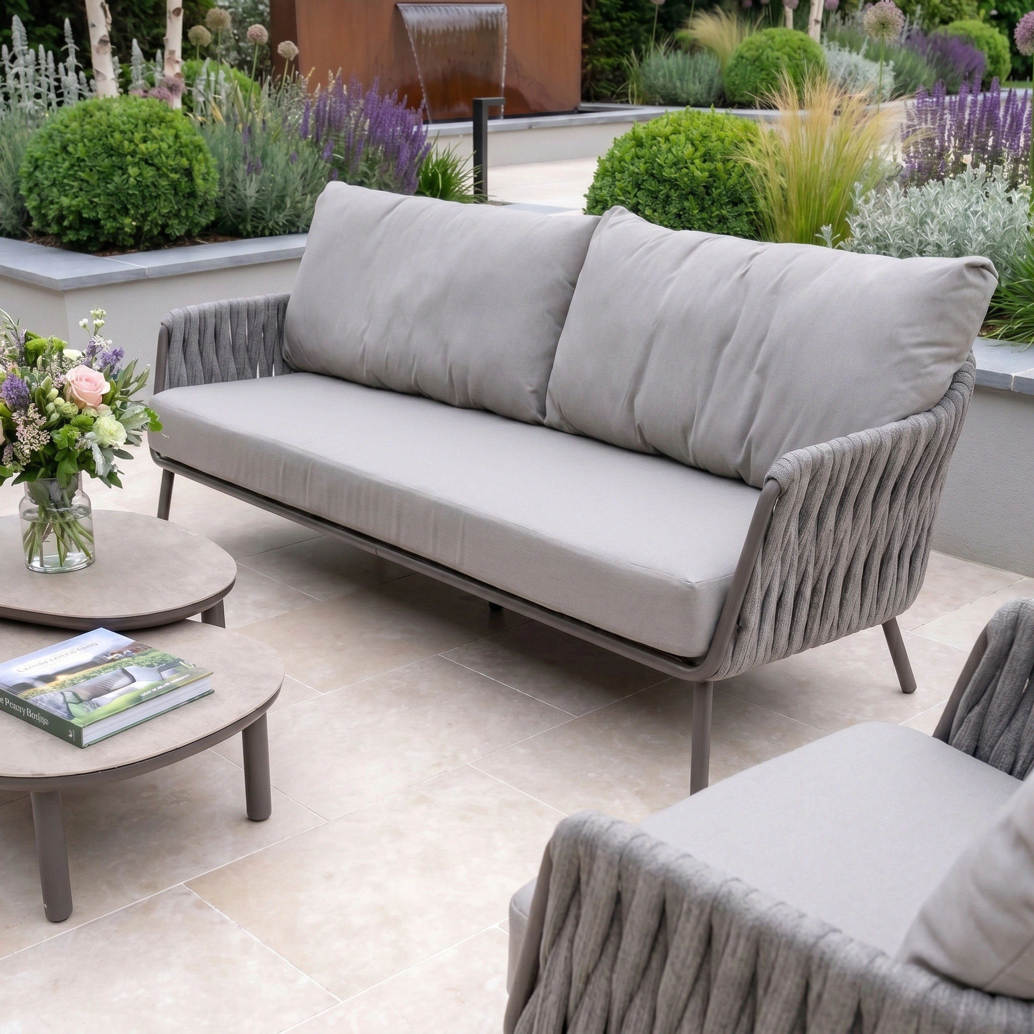 Modern patio with the Capri 3 Seat Sofa Set in Stone Grey, armchair, coffee tables, flowers, and a lush green garden—ideal garden furniture for stylish outdoor relaxation.
