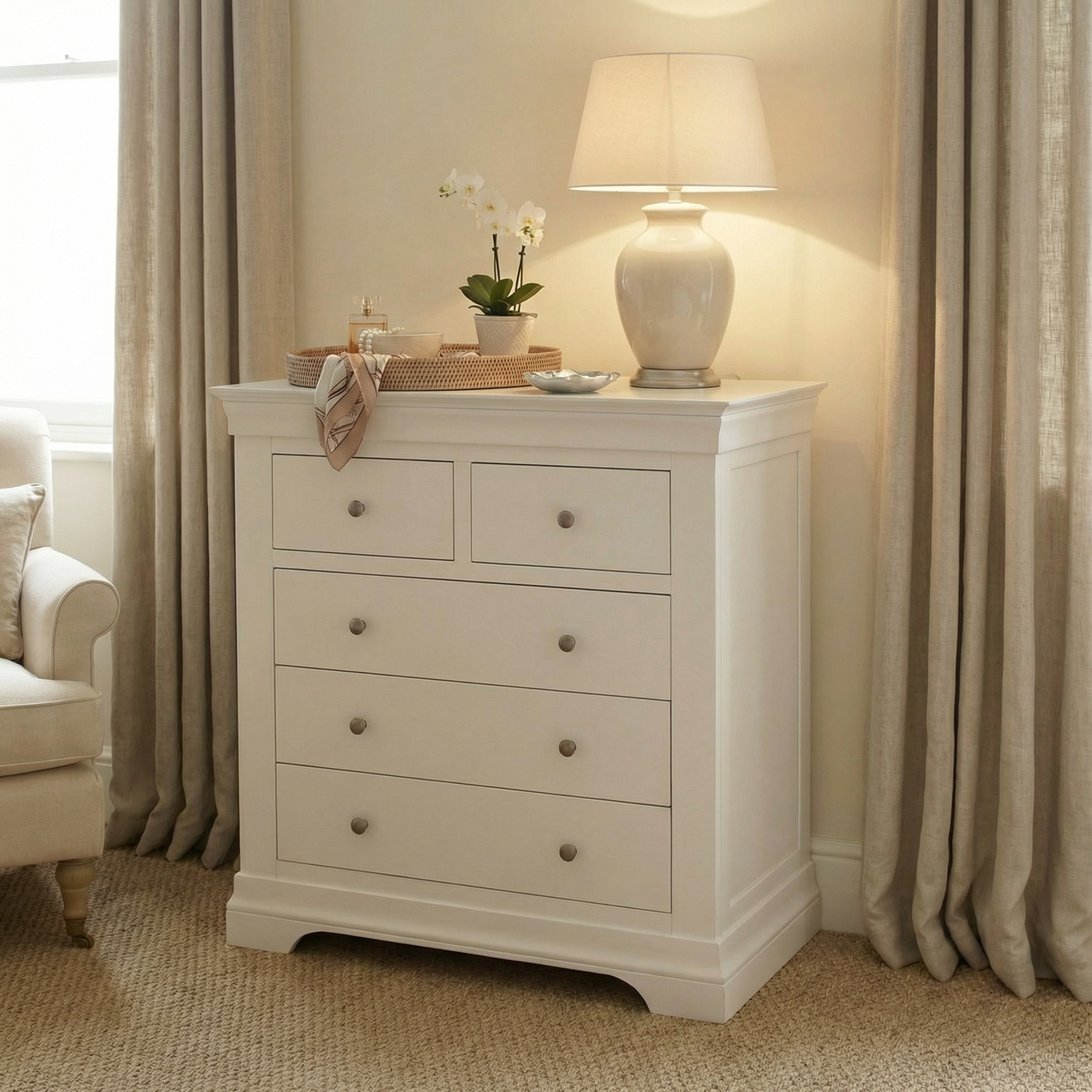 The Chalbury 2 Over 3 Chest of Drawers in Warm White is shown with a lamp, flowers, and basket on top, placed beside beige curtains and an armchair.