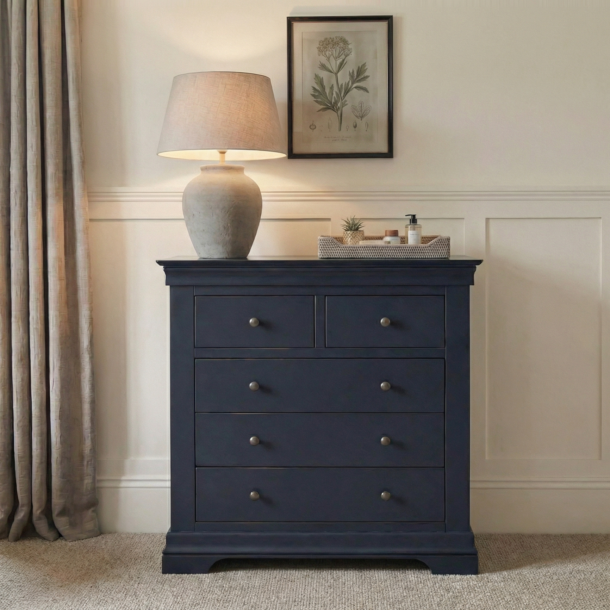 A Chalbury 2 Over 3 Chest of Drawers in Dusky Black sits beneath a lamp and botanical print, next to beige curtains.