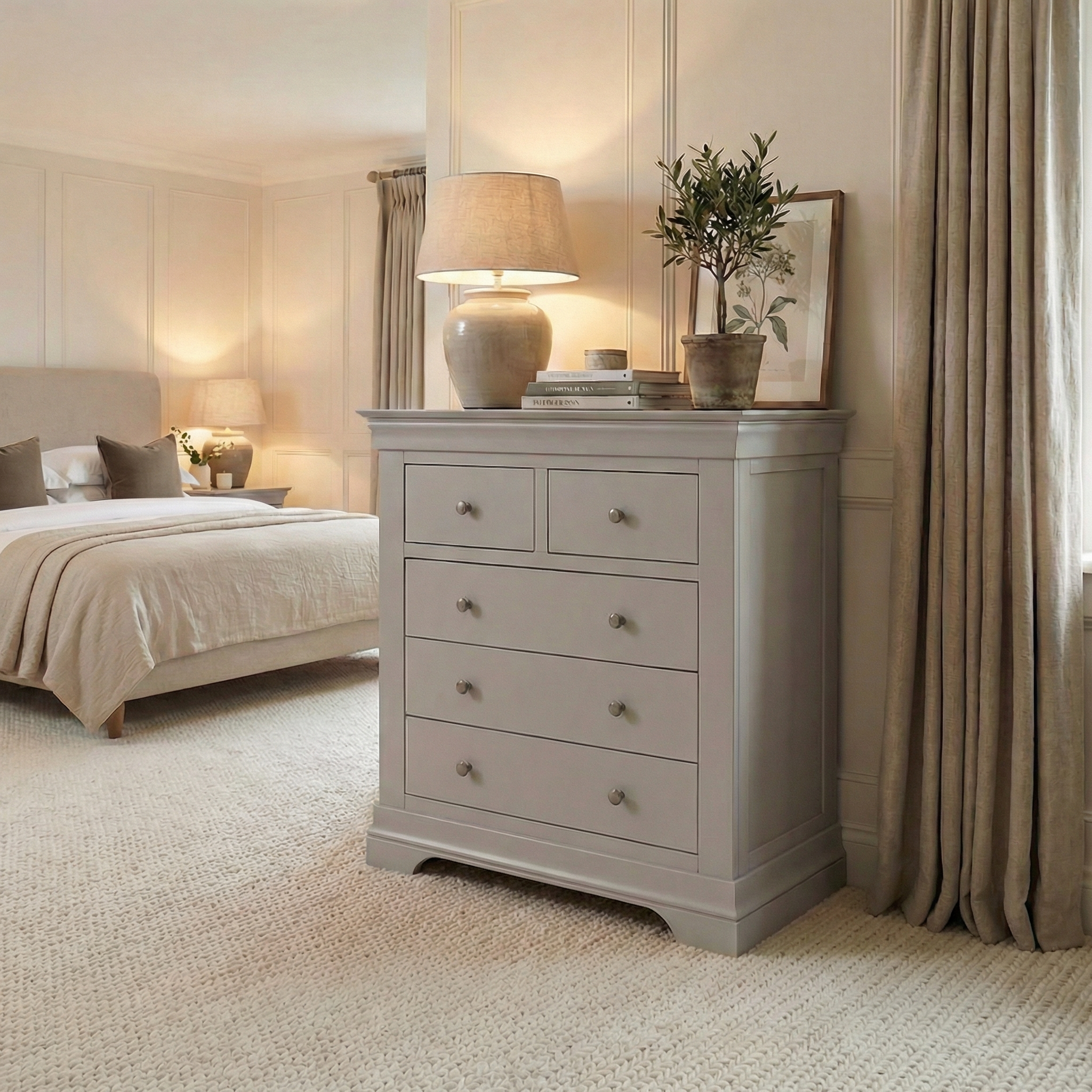A cozy, neutral bedroom showcases the Chalbury 2 Over 3 Chest of Drawers in Pebble Grey, paired with a lamp, a plant, and a neatly made bed in the background.