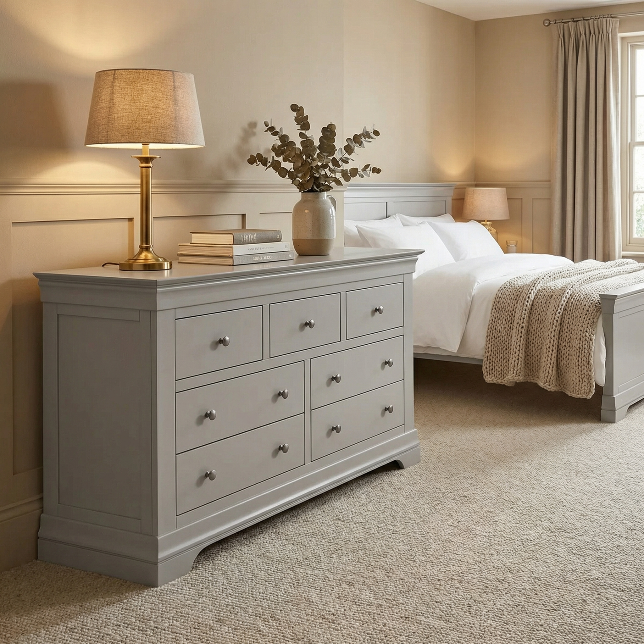 A cozy bedroom with the Chalbury 3 Over 4 Chest of Drawers in Pebble Grey, lamp, vase, books, bed with white linens, and beige walls and carpet—ideal for stylish bedroom storage.