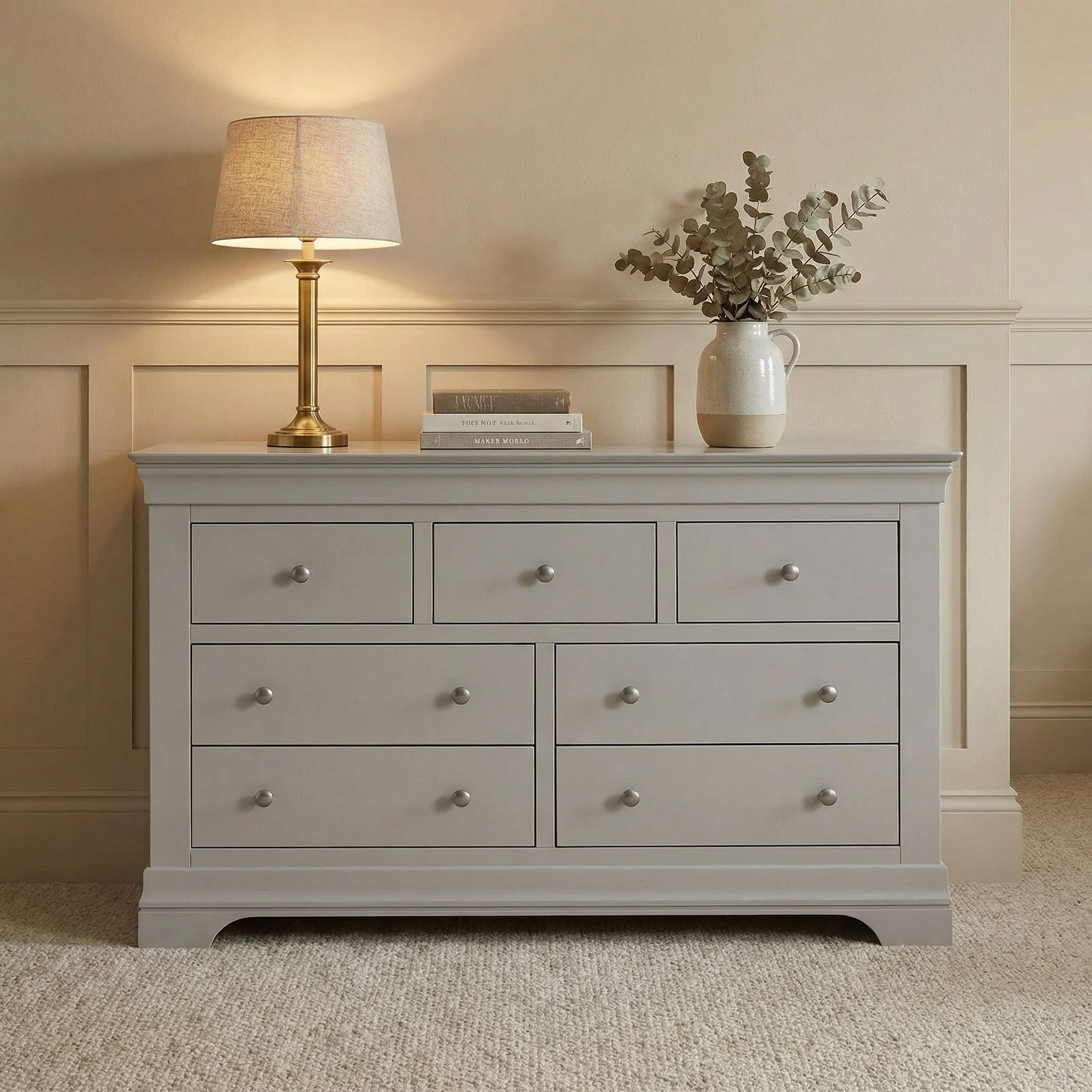 The Chalbury 3 Over 4 Chest of Drawers in Pebble Grey adds stylish storage to your bedroom, featuring spacious drawers and a sleek design, shown with decor accents and set against a beige wall.