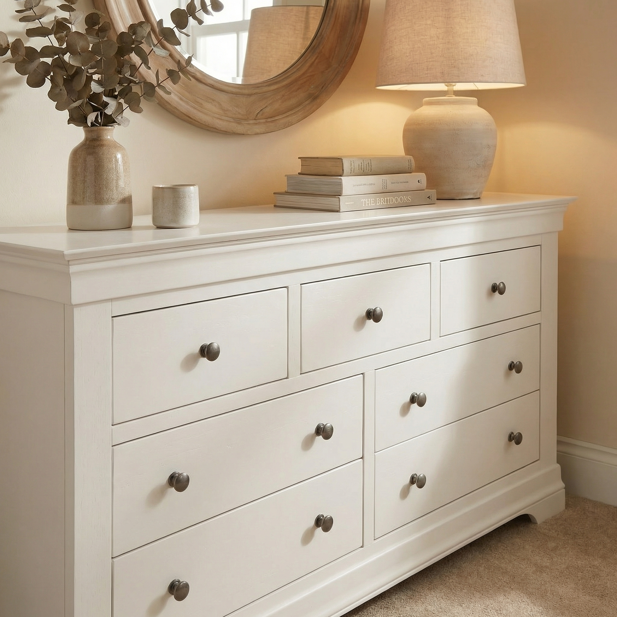 The contemporary Chalbury 3 Over 4 Chest of Drawers in Warm White features seven drawers, styled with a lamp, books, vases on top, and a round mirror mounted above.