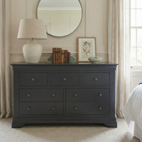 The Chalbury 3 Over 4 Chest of Drawers in Dusky Black sits beneath a round mirror in a bright bedroom, topped with a lamp, books, and framed art.