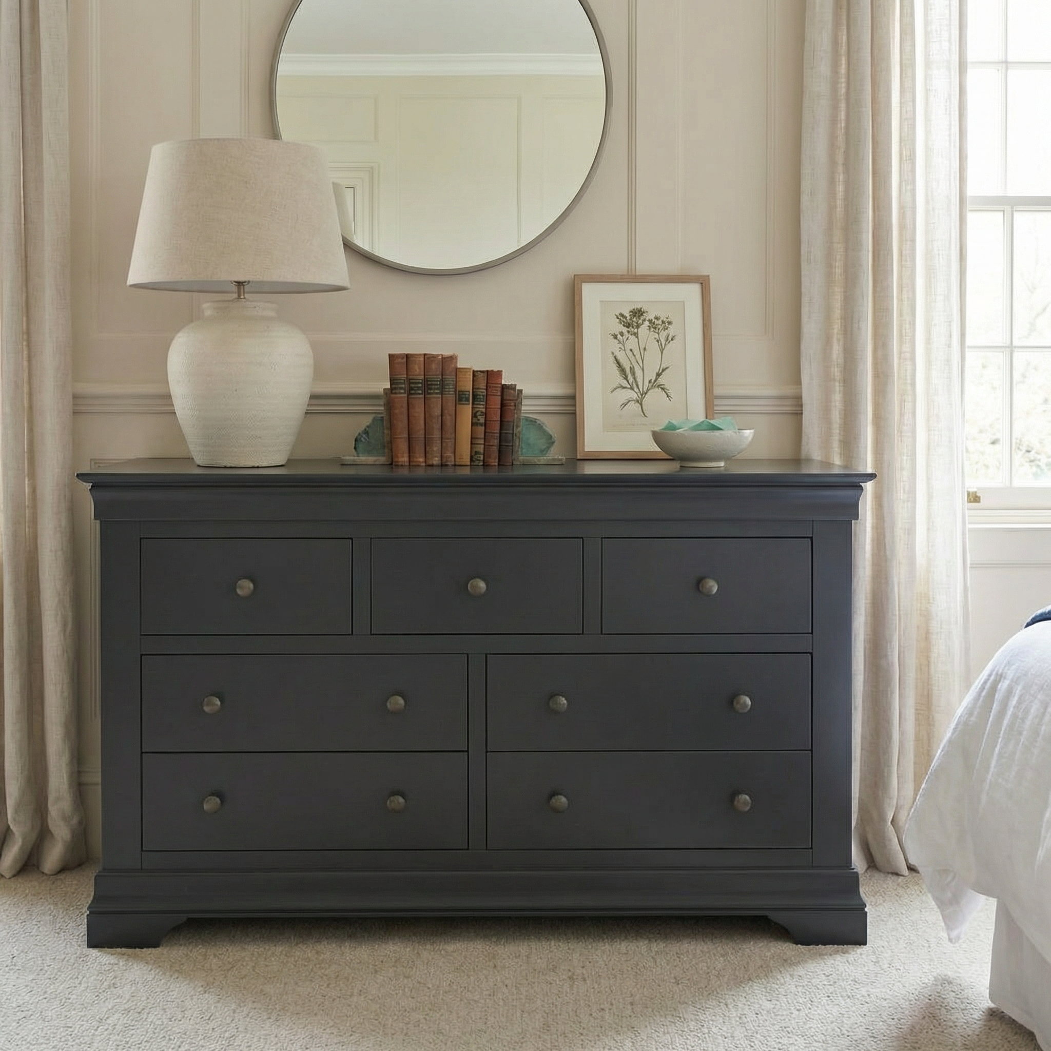 A Chalbury 3 Over 4 Chest of Drawers in Dusky Black styled with a lamp, books, framed art, bowl, and round mirror completes this bright, modern bedroom.