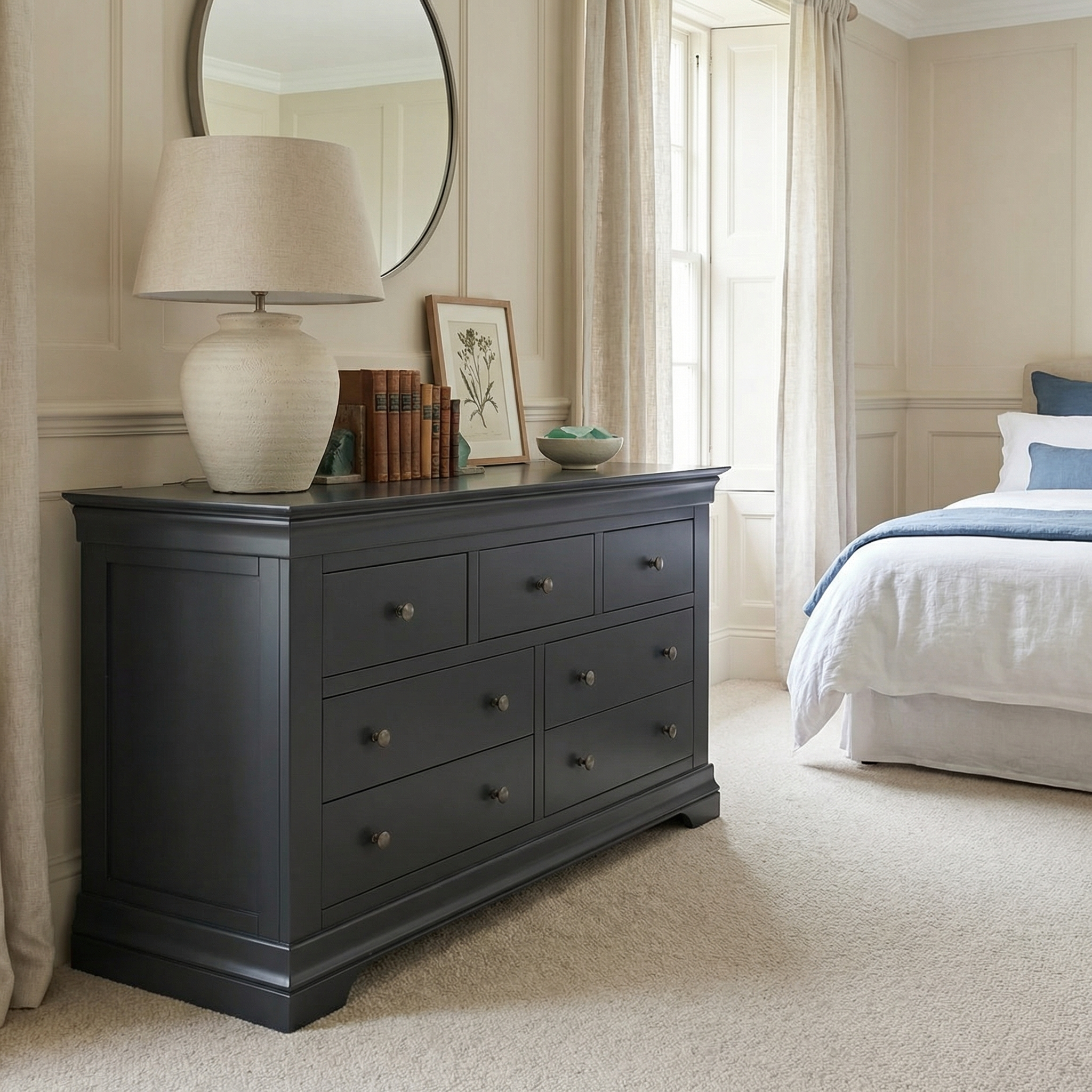 A Chalbury 3 Over 4 Chest of Drawers in Dusky Black, topped with a lamp, books, and art, sits beside the bed in this bright, neutral-toned modern bedroom.