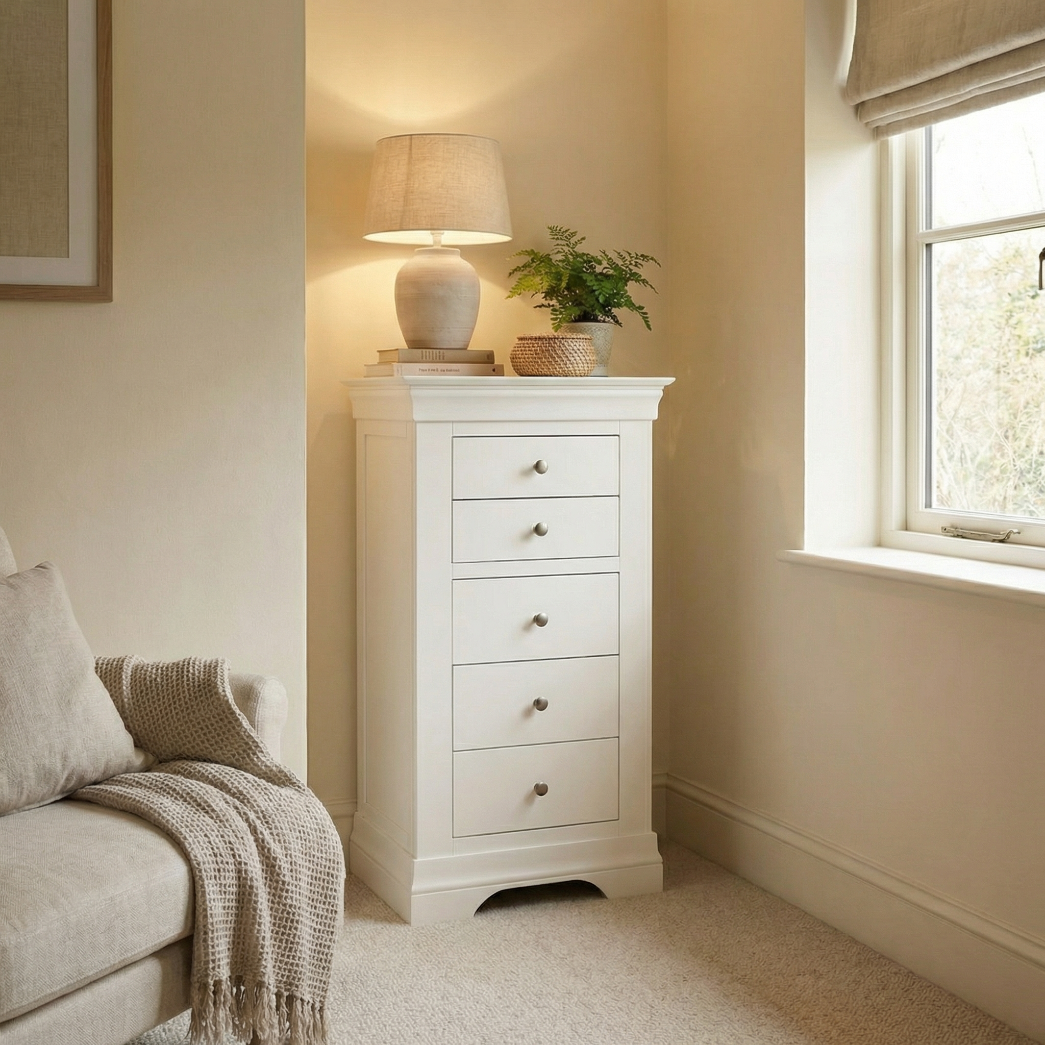 The Chalbury 5 Chest of Drawers in Warm White sits beside a window topped with a lamp, books, and plant—ideal for bedroom storage or adding elegance to any cozy, neutral-toned living room corner.