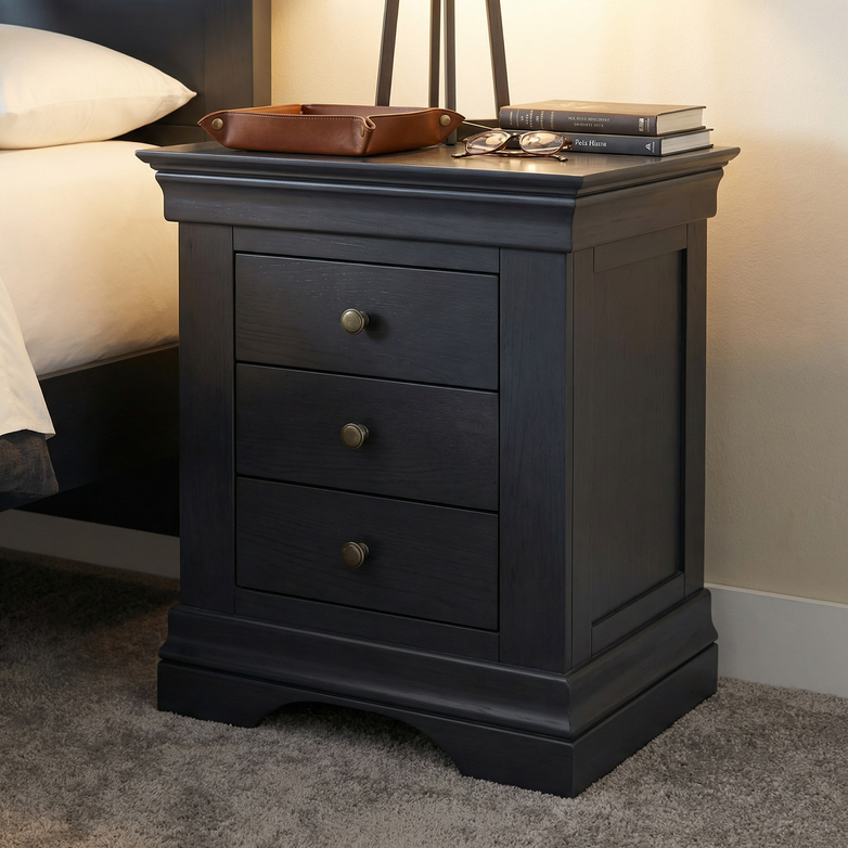 The Chalbury 3 Drawer Bedside Table in Dusky Black offers ample bedroom storage with three drawers, and sits stylishly by the bed, topped with a lamp, books, tray, and glasses.