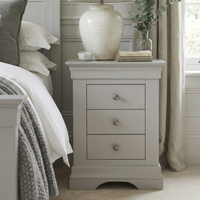 The Chalbury 3 Drawer Bedside Table in Pebble Grey, styled with a lamp, books, and a plant beside a tidy bed, is perfect for bringing modern flair to your room.
