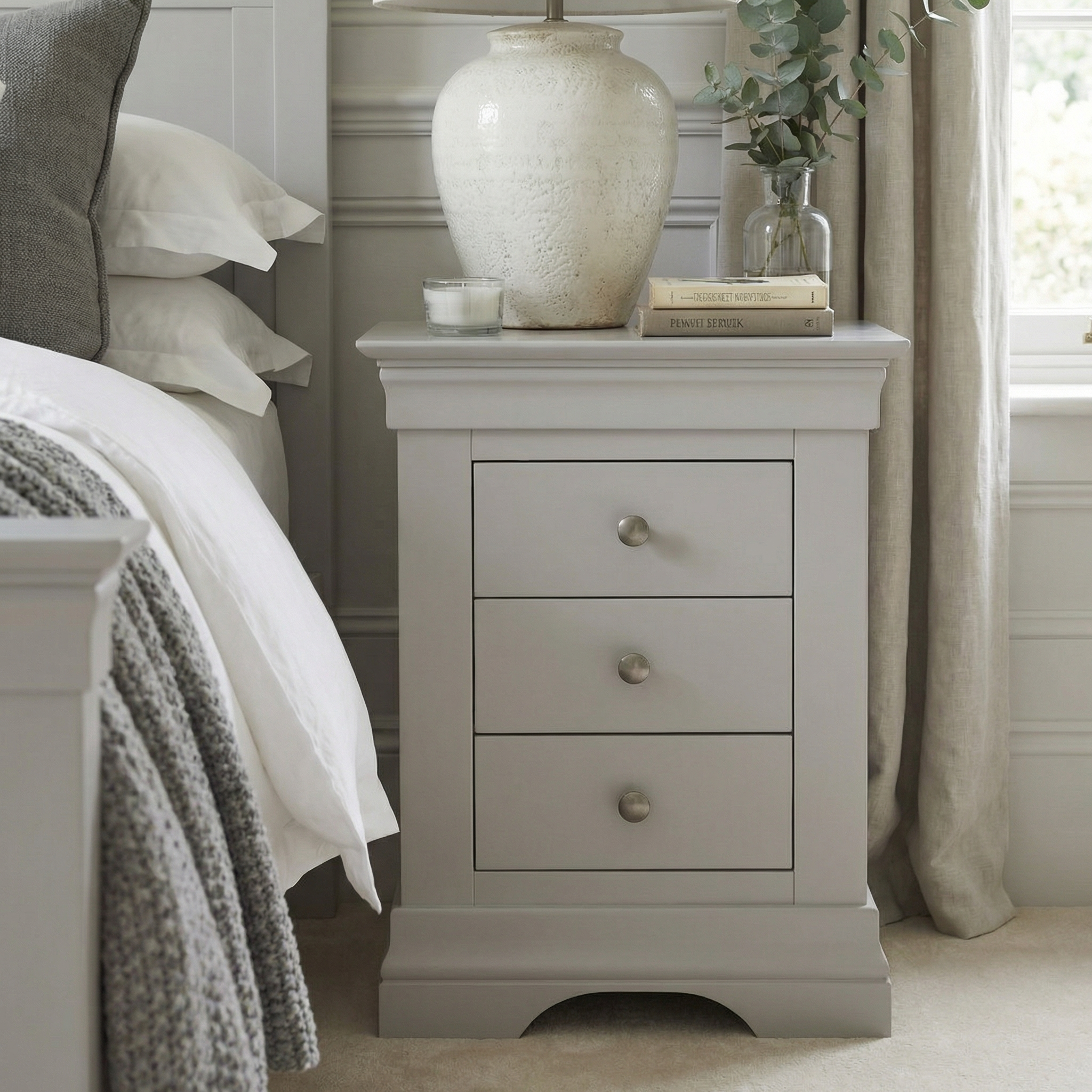 The Chalbury 3 Drawer Bedside Table in Pebble Grey, styled with a lamp, books, and a plant beside a tidy bed, is perfect for bringing modern flair to your room.
